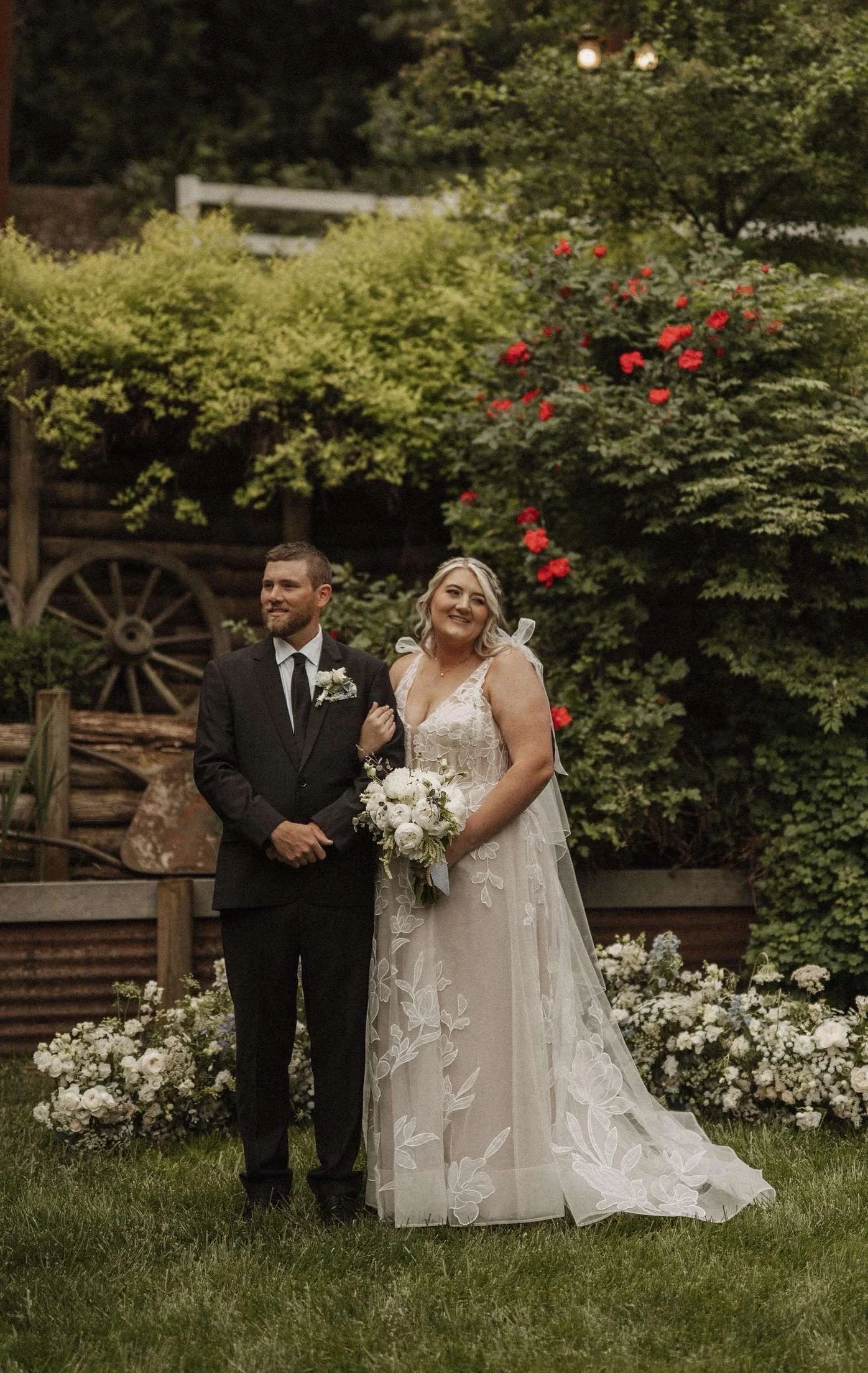 A bride and groom standing in a garden with greenery and flowers, the bride holding a bouquet, both dressed in wedding attire, smiling at the camera.