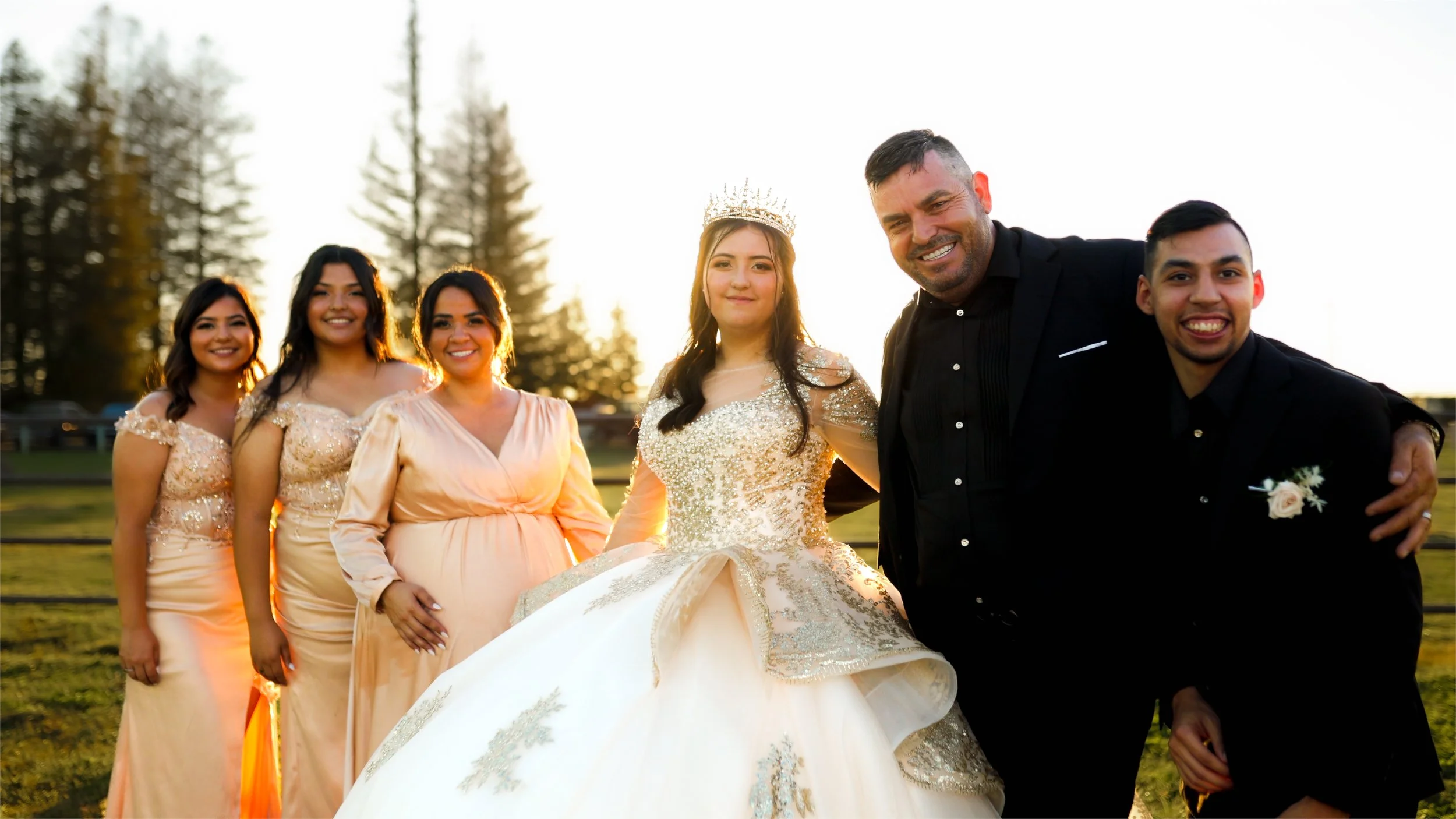 Group of people celebrating outdoors during golden hour, including a woman in a wedding gown wearing a crown, and others in formal attire, smiling at the camera.