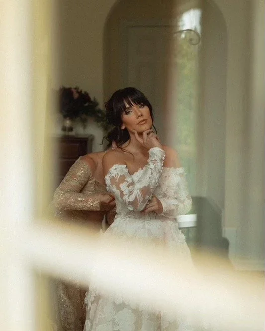 A woman in a white wedding dress with lace details, posing thoughtfully, with a reflection visible in a mirror.