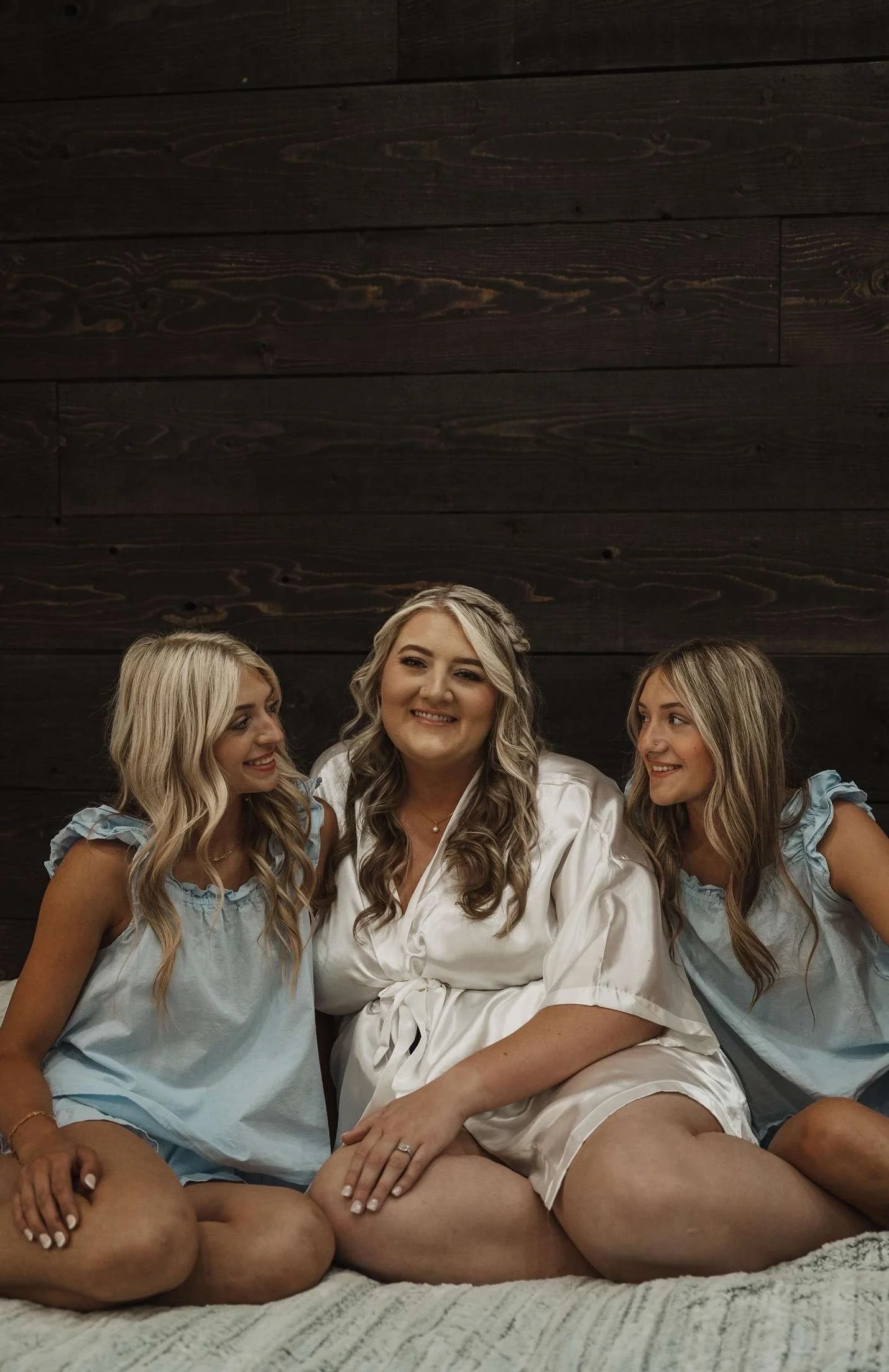 Three women sitting on a bed, smiling, with a dark wooden wall behind them. They are wearing light-colored sleepwear.