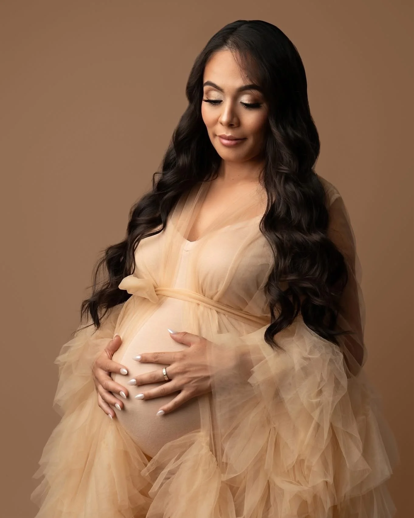 A pregnant woman with long dark hair wearing a flowing beige dress, gently holding her belly and looking down.