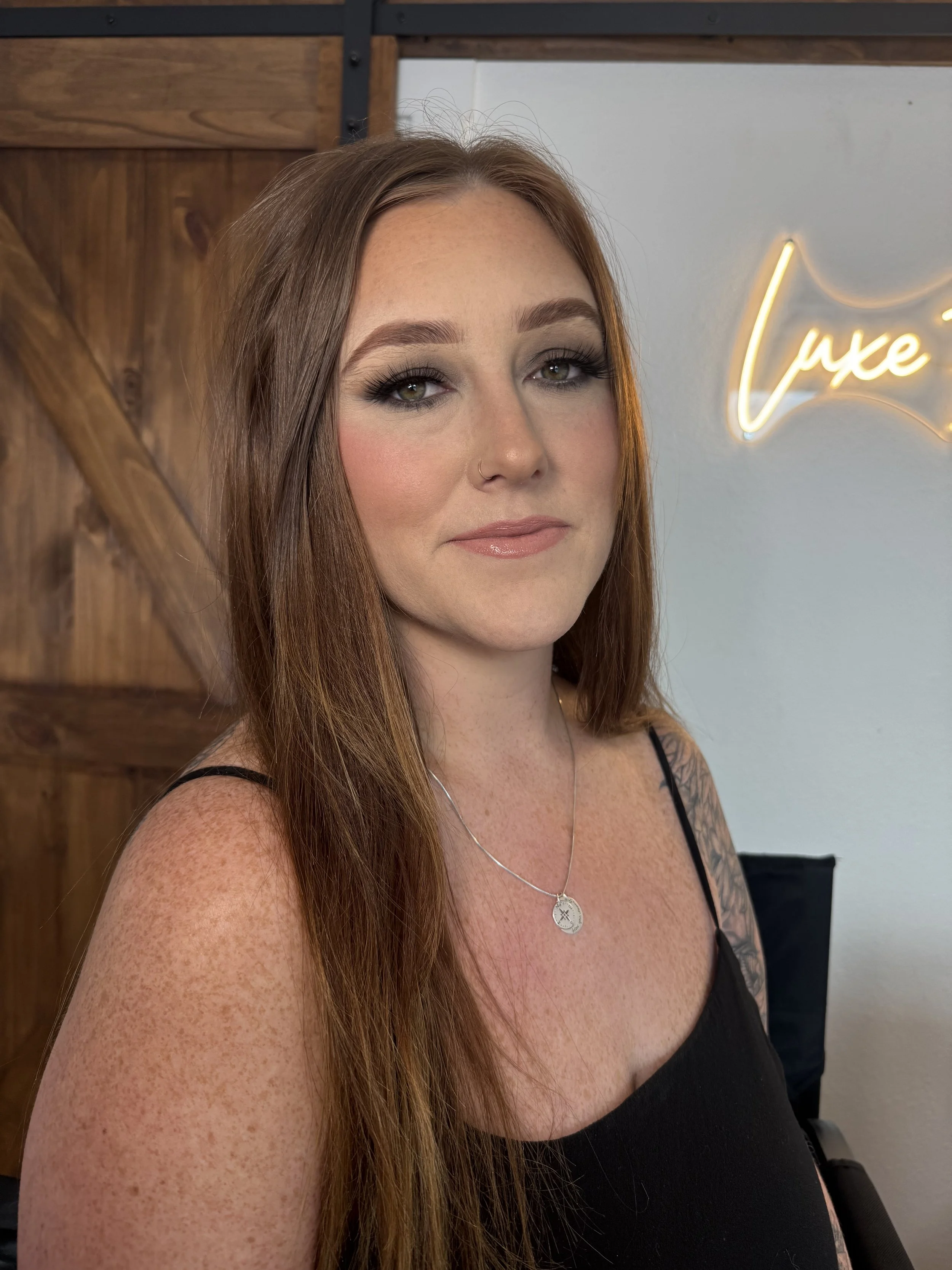 A woman with long red hair, wearing a black tank top and a necklace with a circular pendant, posing indoors with wood paneling and a neon sign reading 'luxe' in the background.