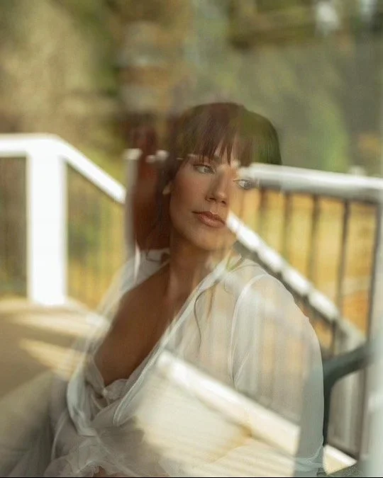 A woman with dark hair and bangs sitting on a porch, looking out through a glass window with reflections.