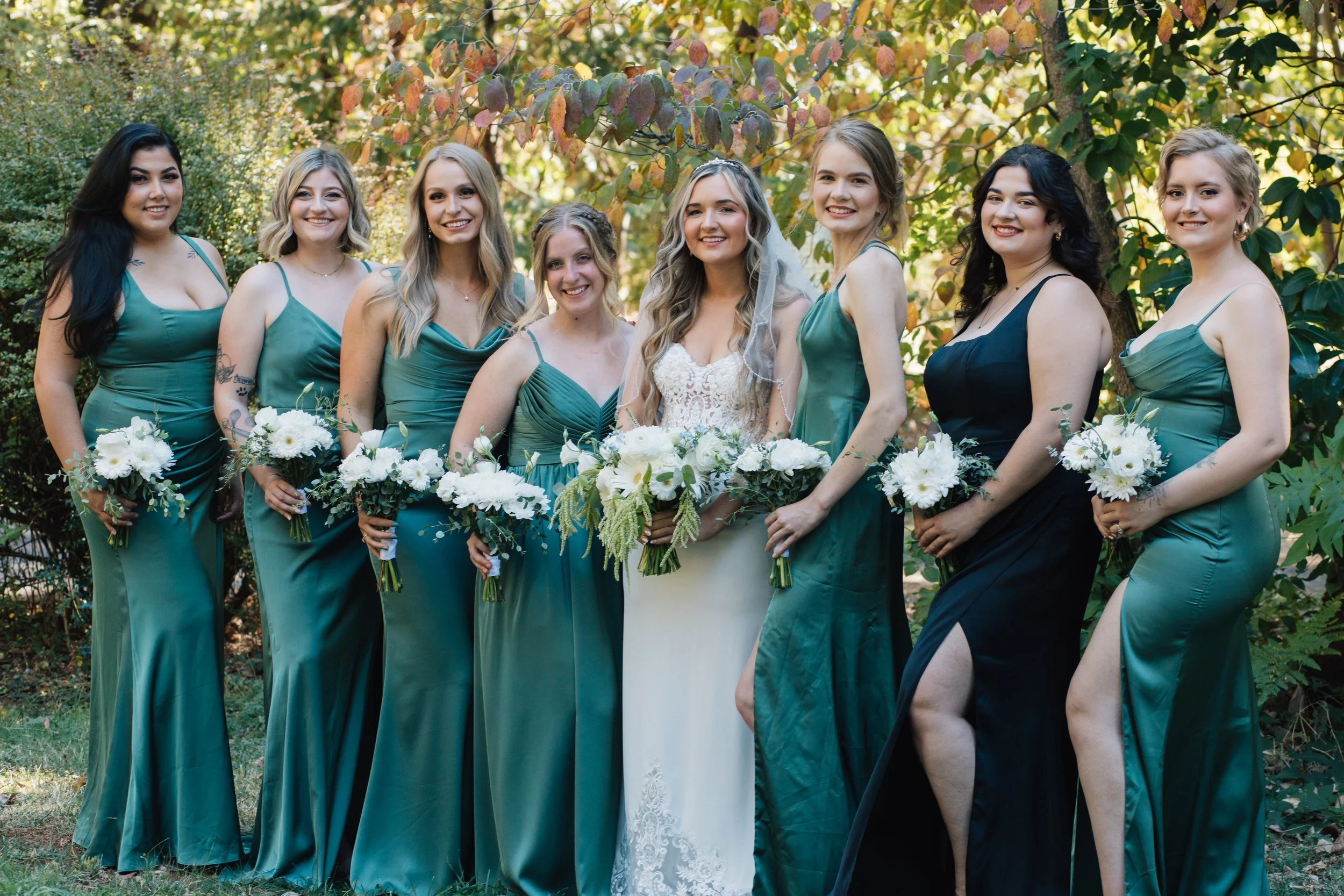 A bride in a white wedding dress surrounded by eight bridesmaids in teal and black dresses holding bouquets of white flowers outdoors.