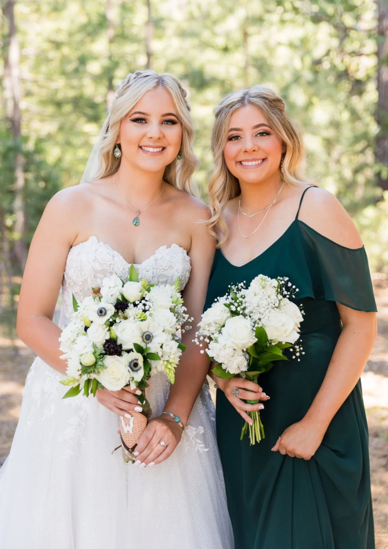 Two women standing outdoors in a forested area, dressed for a wedding, holding bouquets of white flowers, smiling at the camera.