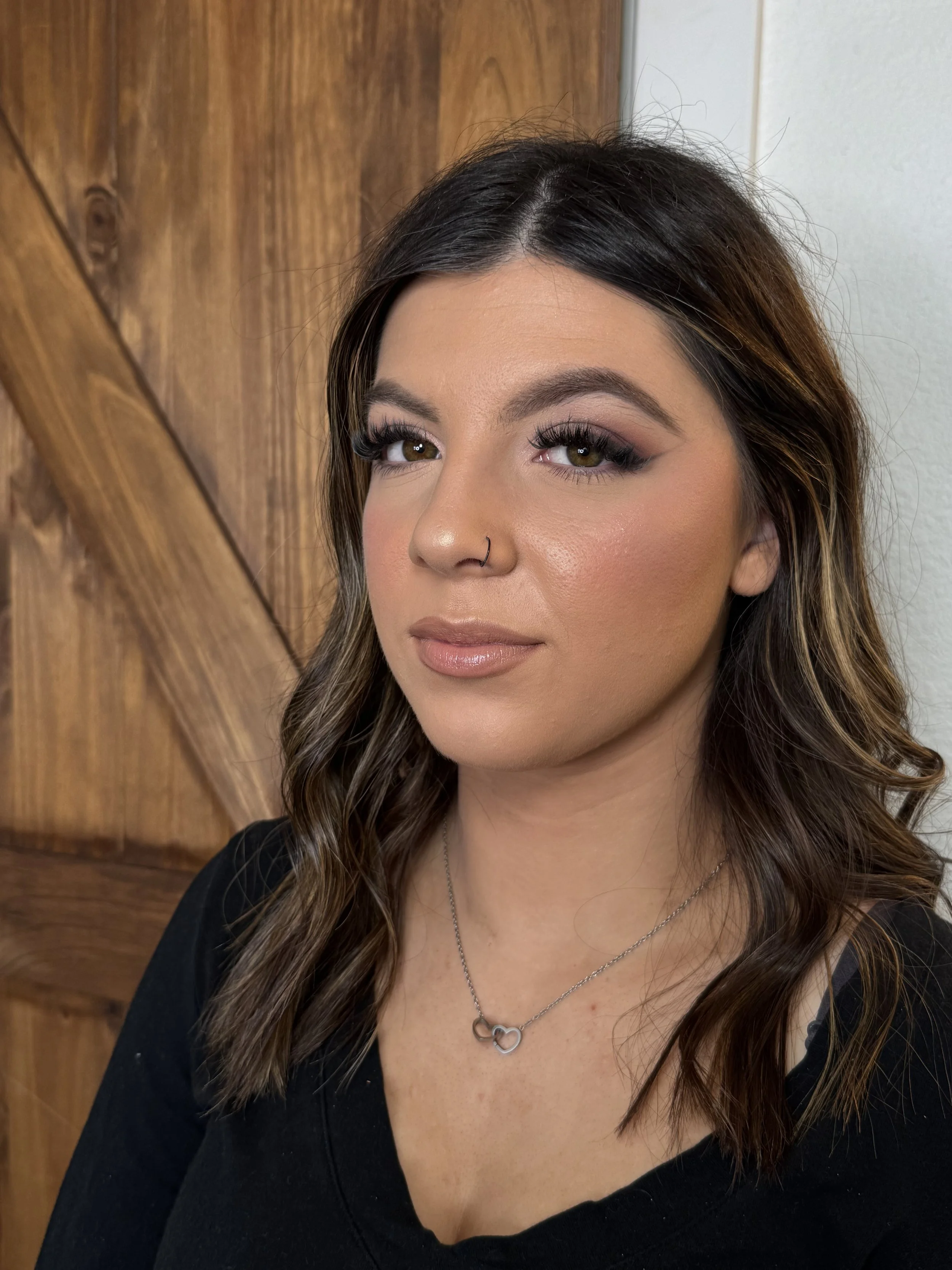 Close-up of a woman with makeup, wavy brown hair with highlights, wearing a black top and a silver necklace with intertwined hearts, standing against a wooden background.
