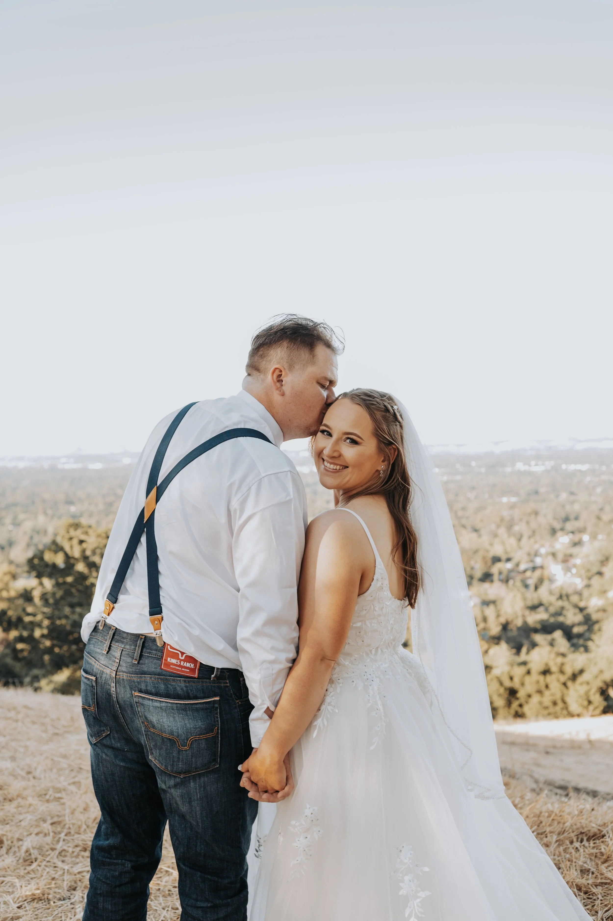 A couple dressed in wedding attire holding hands outdoors, with the groom kissing the bride on the forehead and both smiling. The bride is in a white wedding gown and veil, and the groom in a white shirt with suspenders.