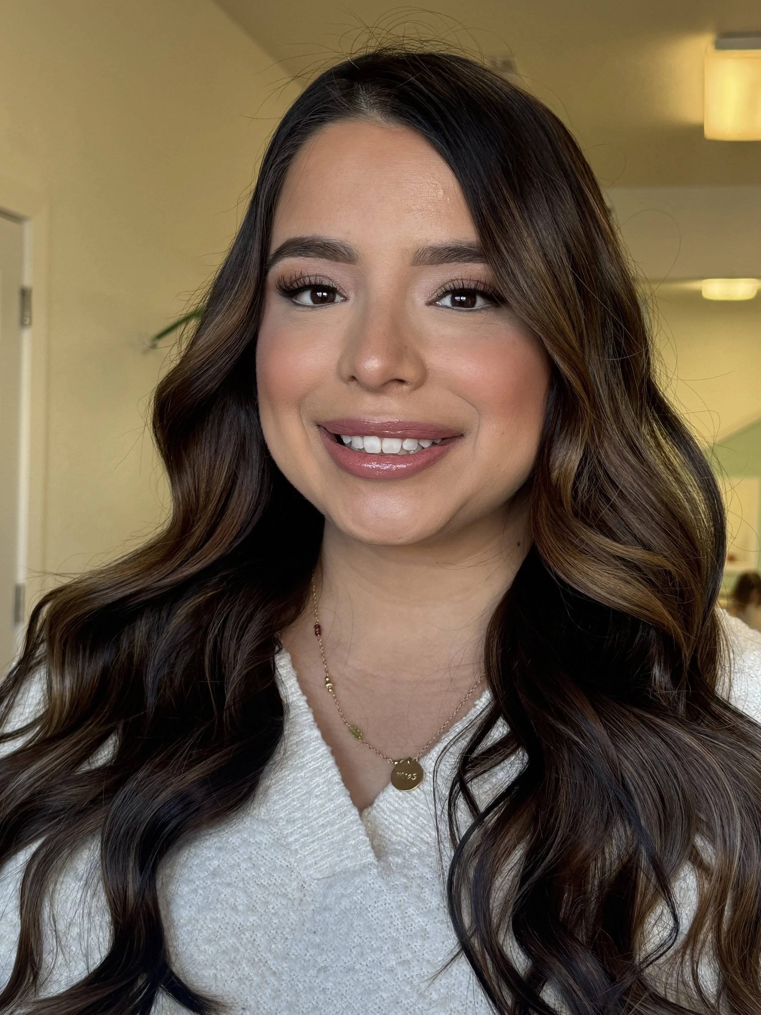 Close-up of a woman with long wavy brown hair, wearing a white top, gold necklace, and smiling at the camera inside a well-lit room.