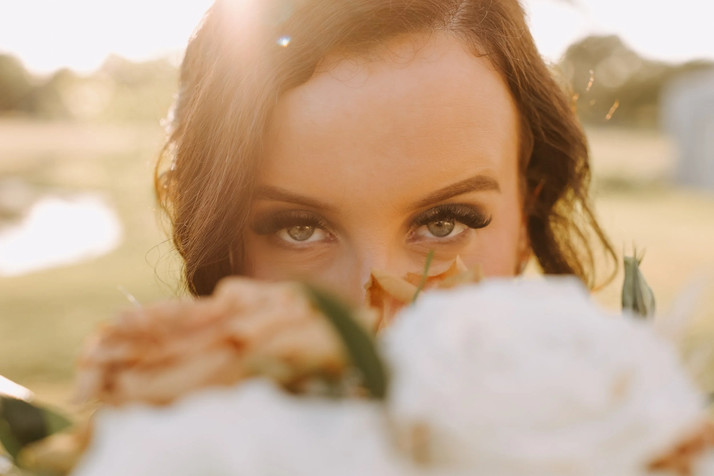 A woman with light brown hair and striking eyes peeks over a fluffy white flower, with a blurred outdoor background and sunlight creating a warm glow.