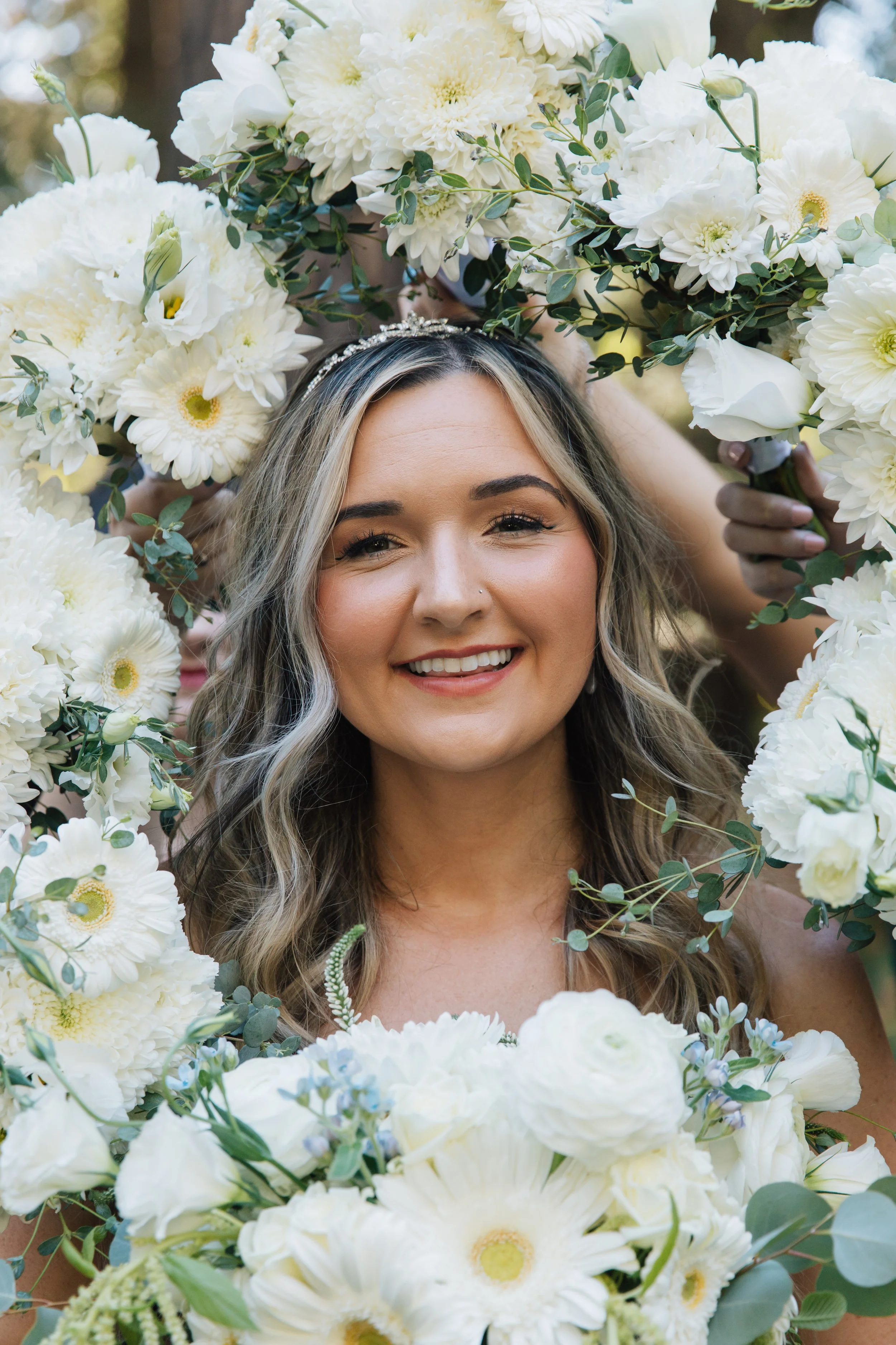 Smiling bride with wavy hair and a tiara, holding a large floral wreath of white flowers and greenery.