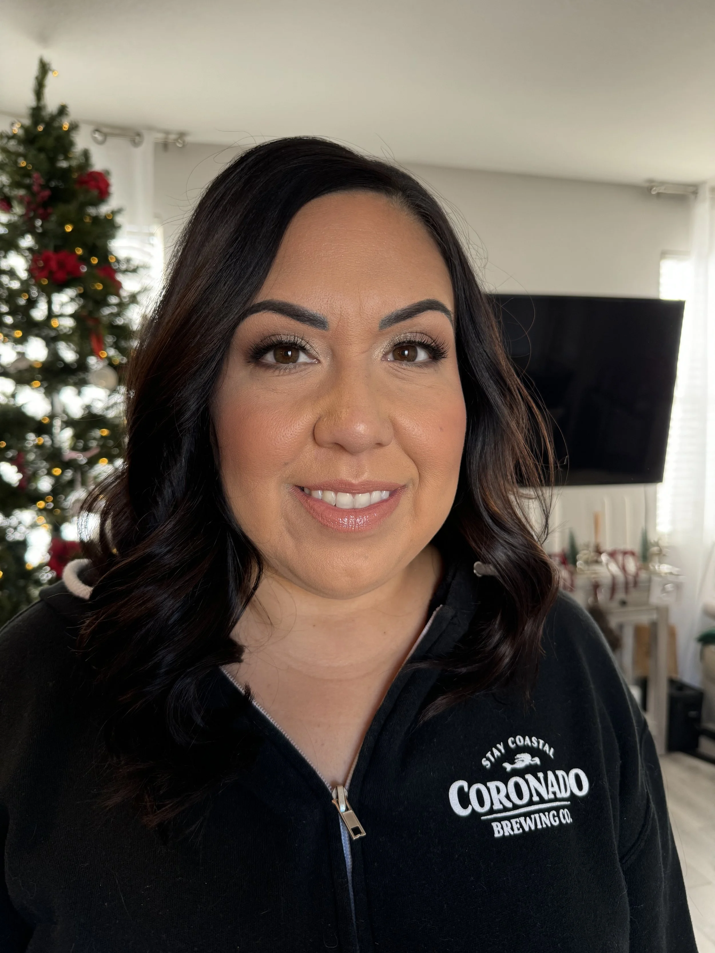 A woman with dark brown, wavy hair smiling in a living room with a decorated Christmas tree in the background, wearing a black zip-up jacket with 'Stay Coastal Coronado Brewing Co.' logo.