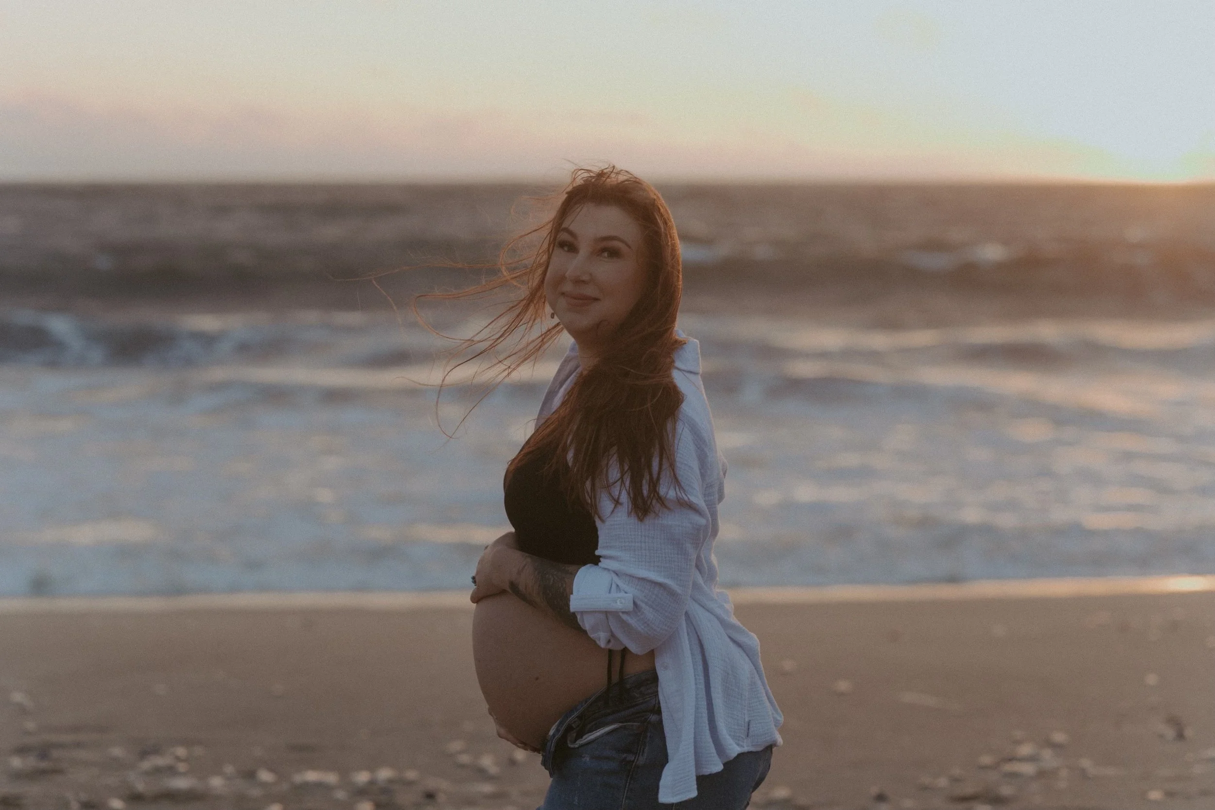 A pregnant woman with long red hair standing on a beach at sunset, smiling, with the ocean and sky behind her.