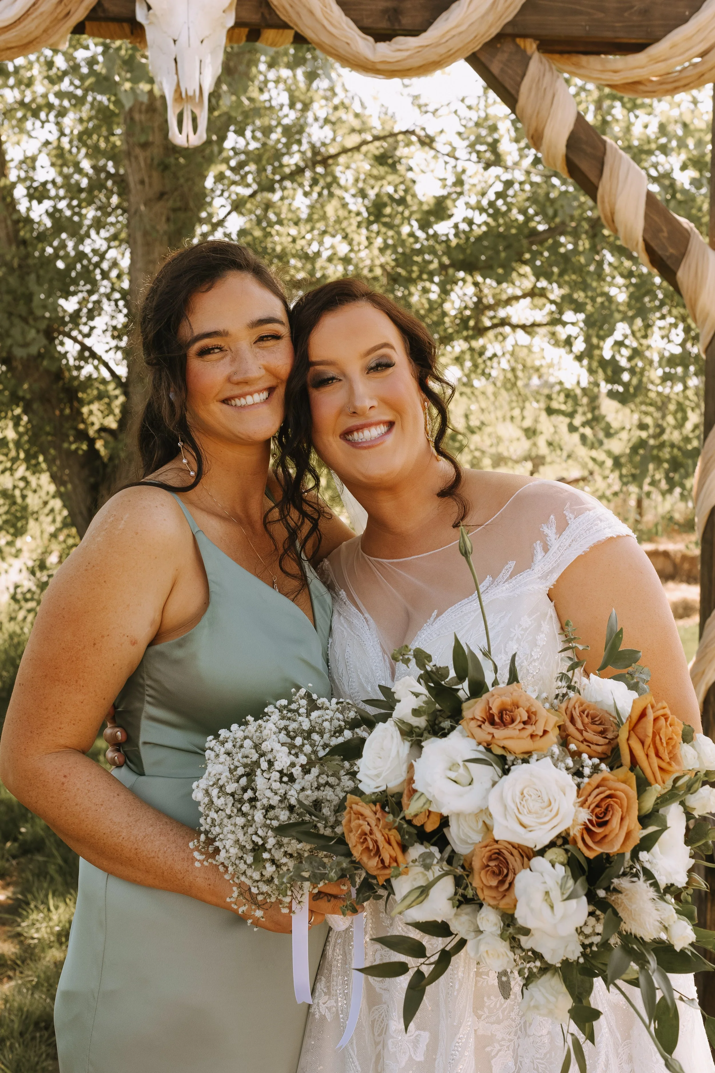 Two women smiling and standing close together outdoors, one is in a wedding dress holding a bouquet of flowers, the other woman is in a light green dress, under a decorated wooden arch.