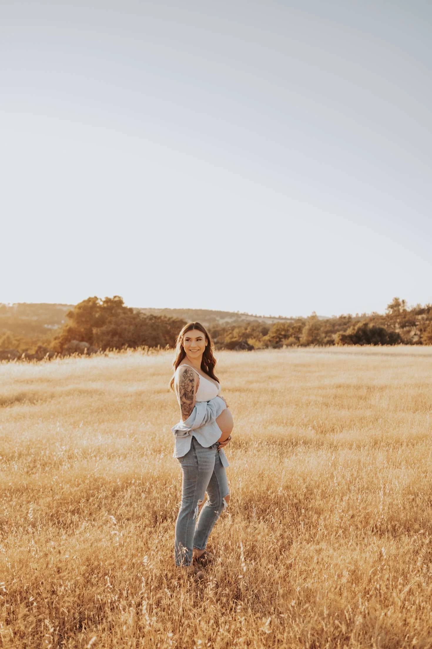 A pregnant woman standing in a field of tall, golden grass during sunset, smiling, with rolling hills and trees in the background.