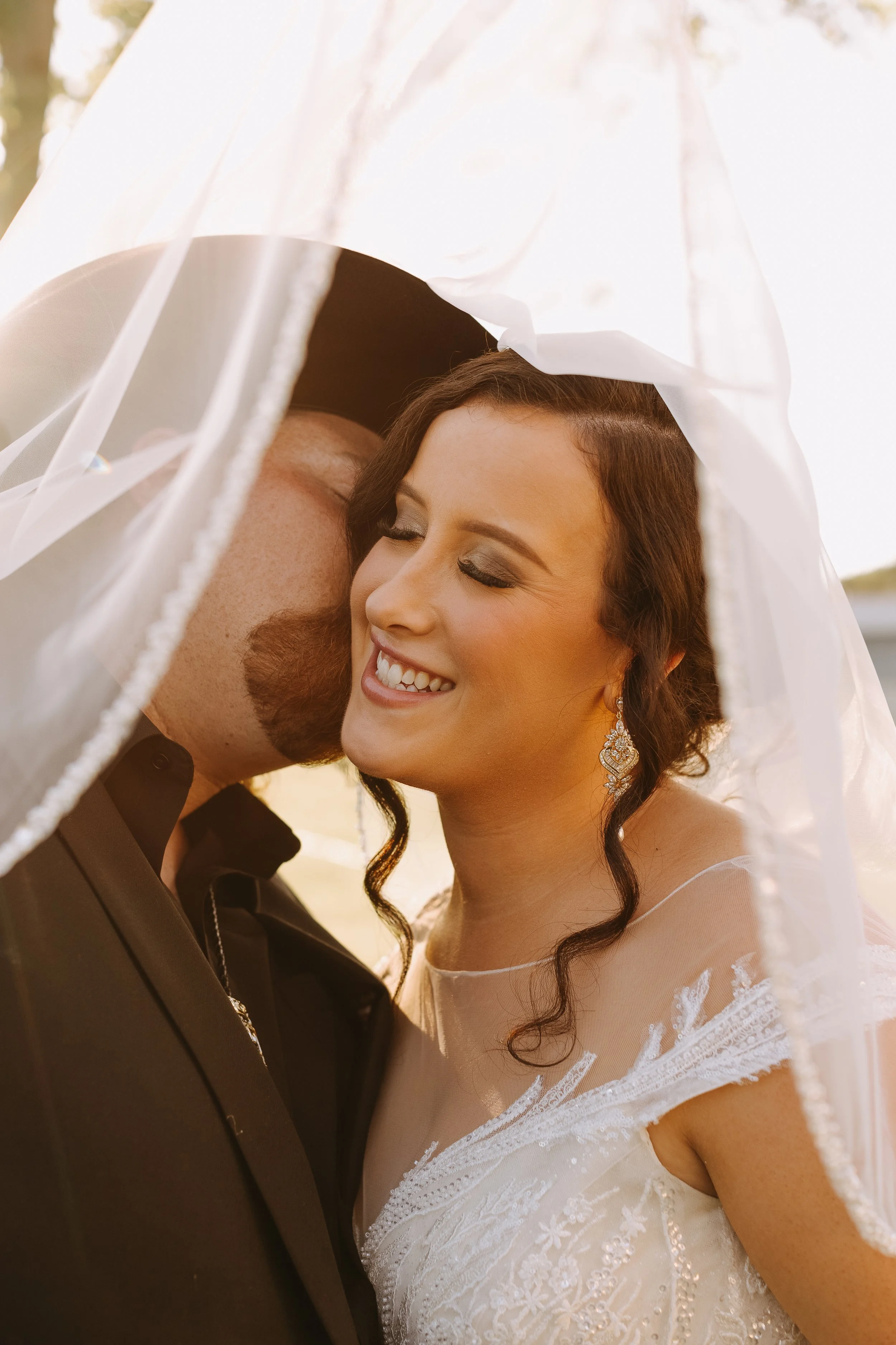 A bride and groom sharing a kiss under a white umbrella on their wedding day, with the bride smiling and wearing a wedding dress and earrings.