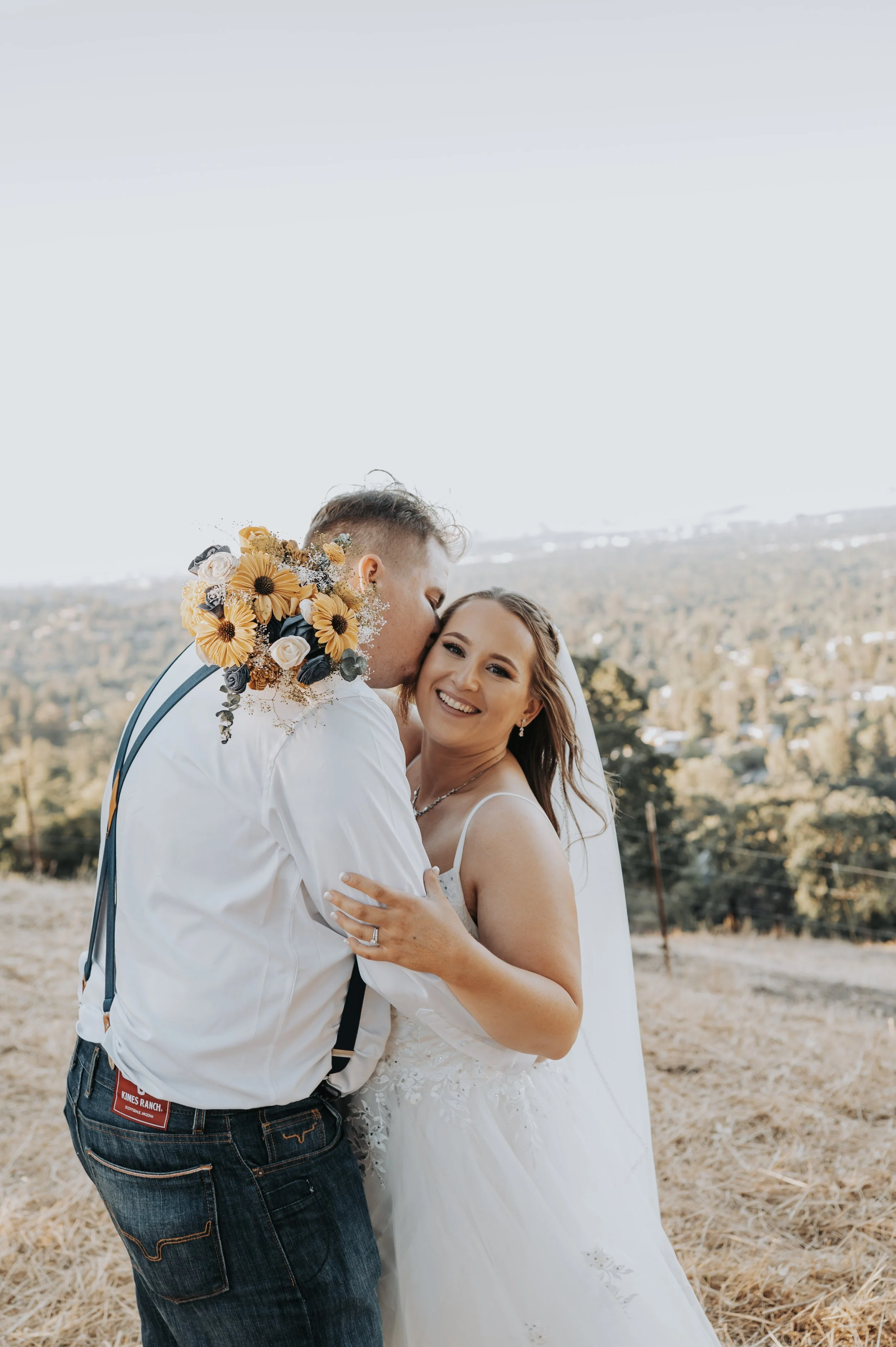 A newlywed couple sharing a kiss outdoors, with the groom holding a bouquet of sunflowers and the bride smiling.