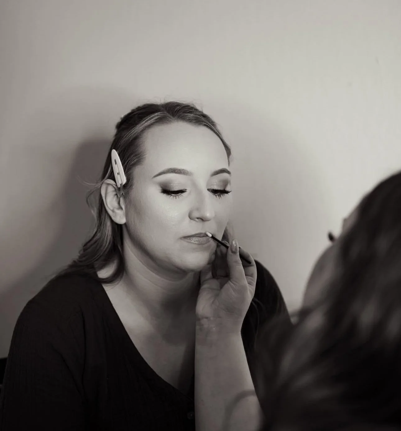 A woman with a hair clip has her makeup applied, with her eyes closed gracefully, by a makeup artist.