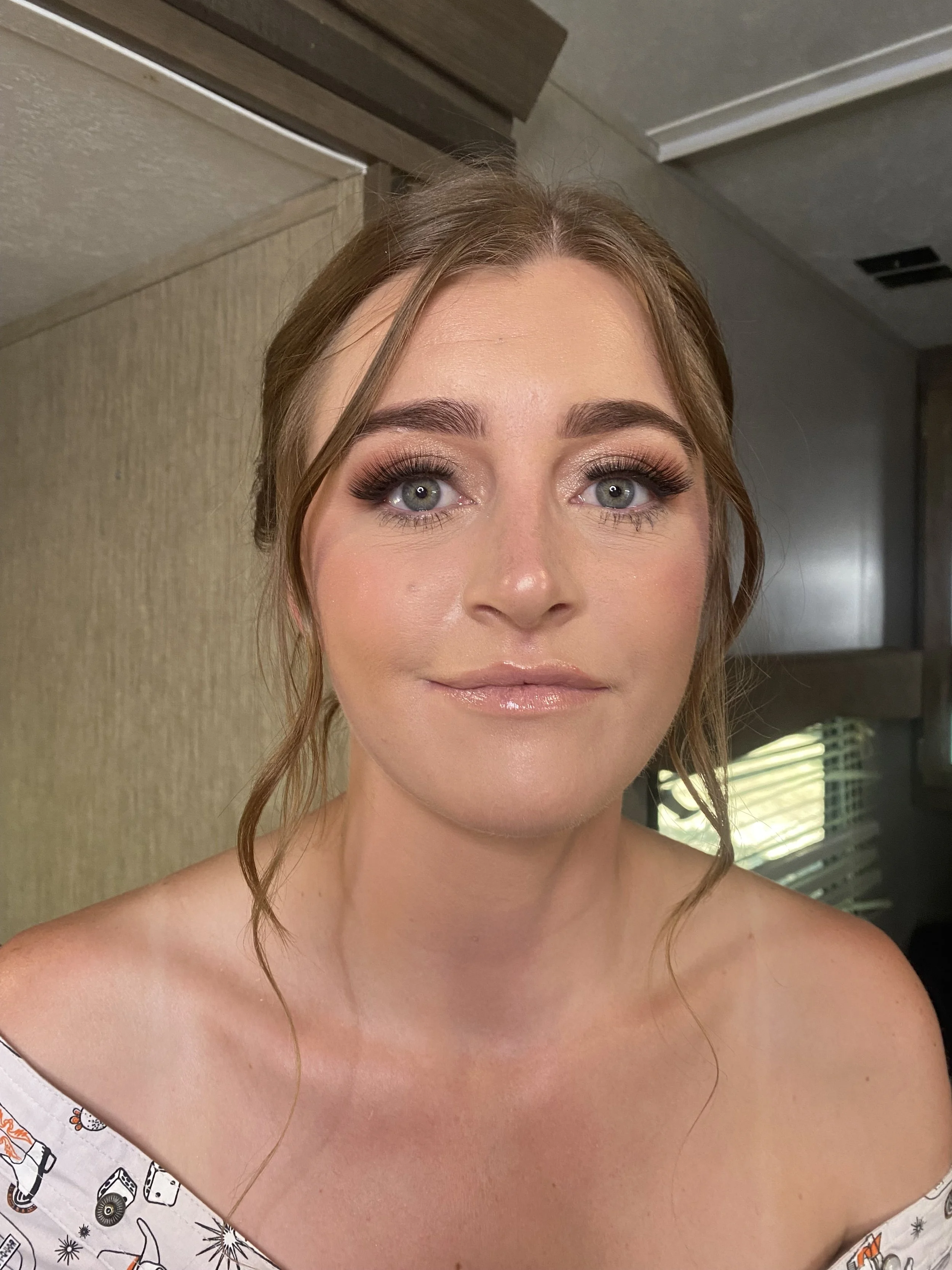 A young woman with blue eyes and light brown hair styled in loose waves, wearing makeup with dark eyeshadow, eyeliner, and nude lipstick. She wears an off-the-shoulder top with a pattern, standing indoors with a neutral-colored wall and a window with