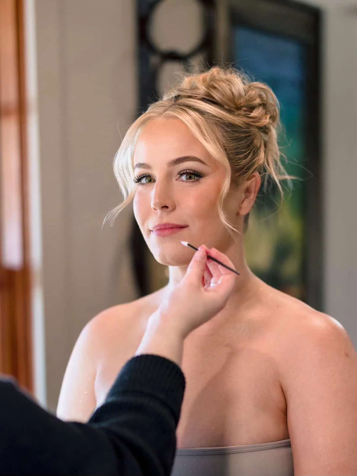A woman with blonde hair styled in an updo getting her makeup done by a makeup artist.