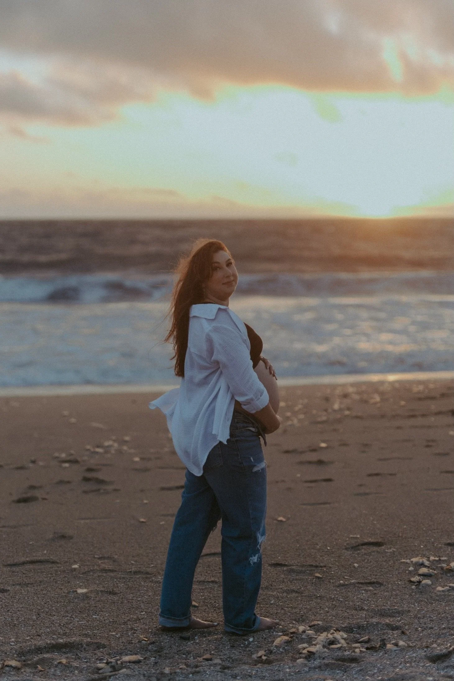 A woman standing on a beach at sunset, cradling her pregnant belly and looking over her shoulder.