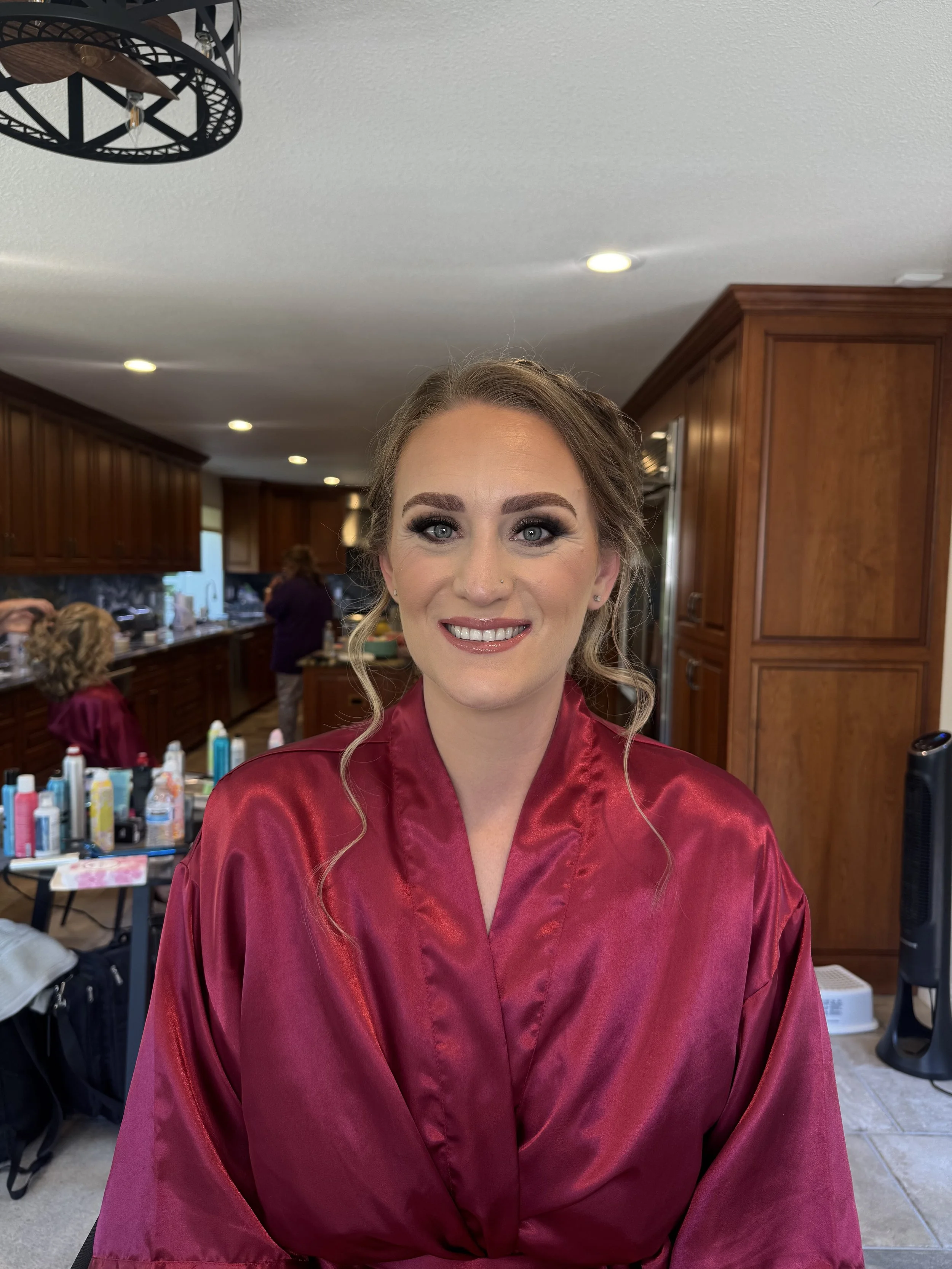 A woman dressed in a burgundy satin robe, smiling, with wedding makeup and hair styled, in a kitchen with wooden cabinets, and makeup products on a table in the background.