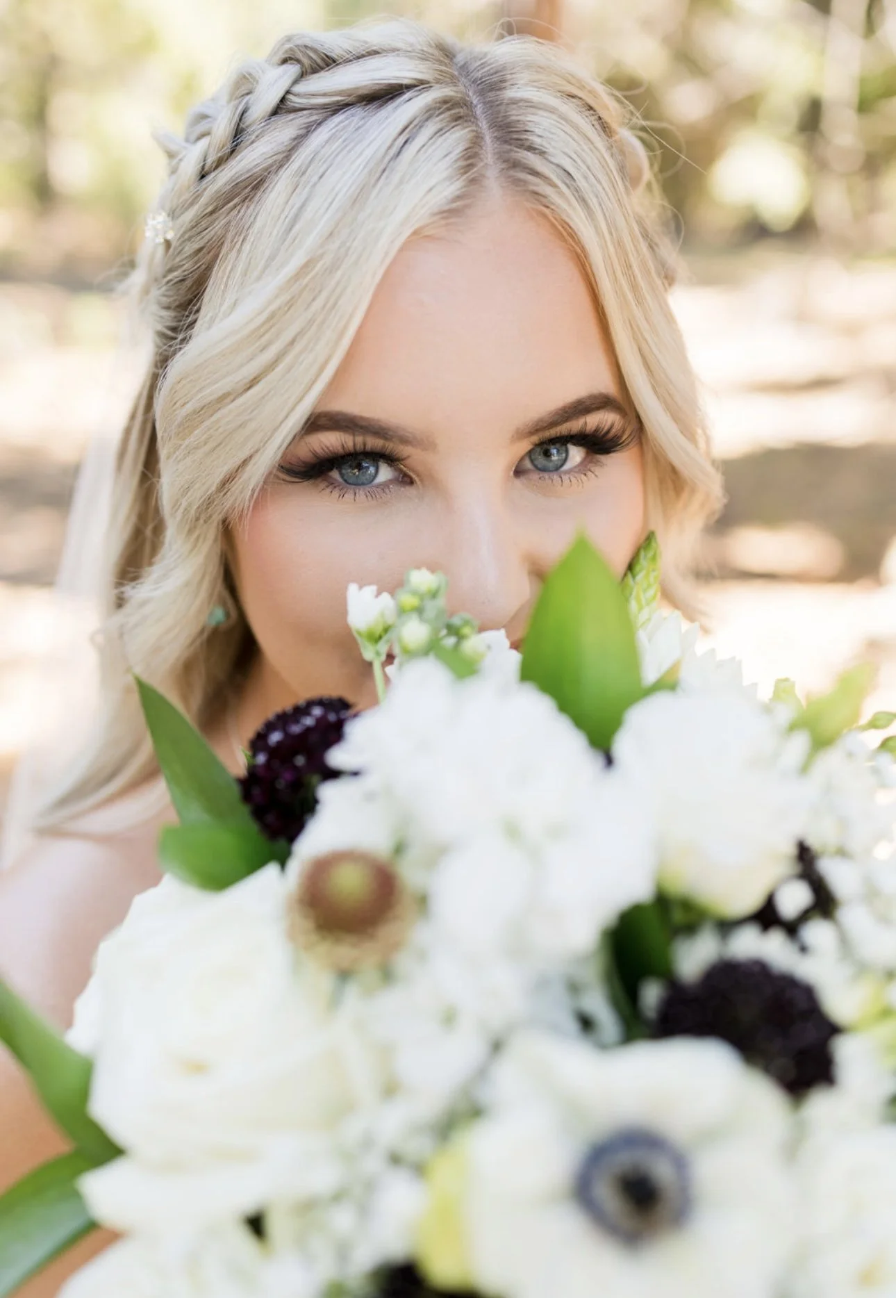 A woman with blonde hair and blue eyes holding a bouquet of white flowers close to her face, outdoors in a natural setting.