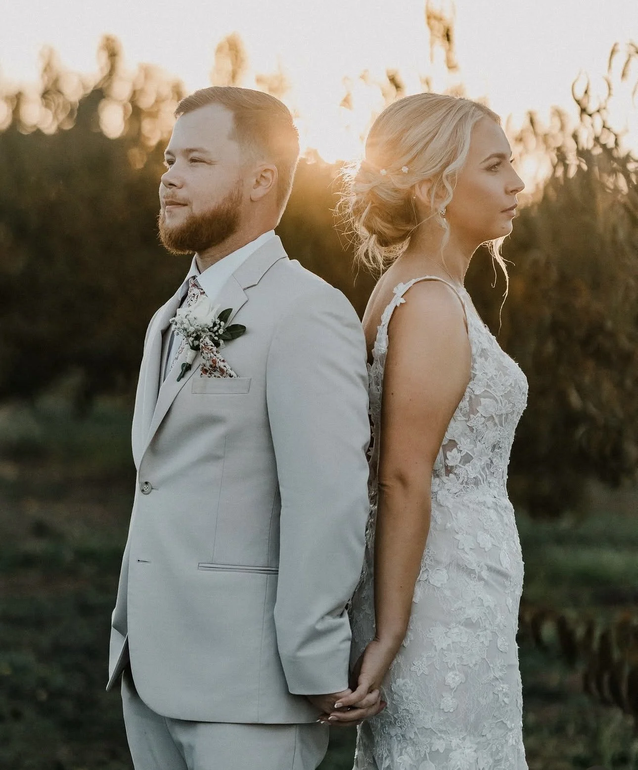 A bride and groom stand back to back outdoors during sunset, holding hands behind their backs.