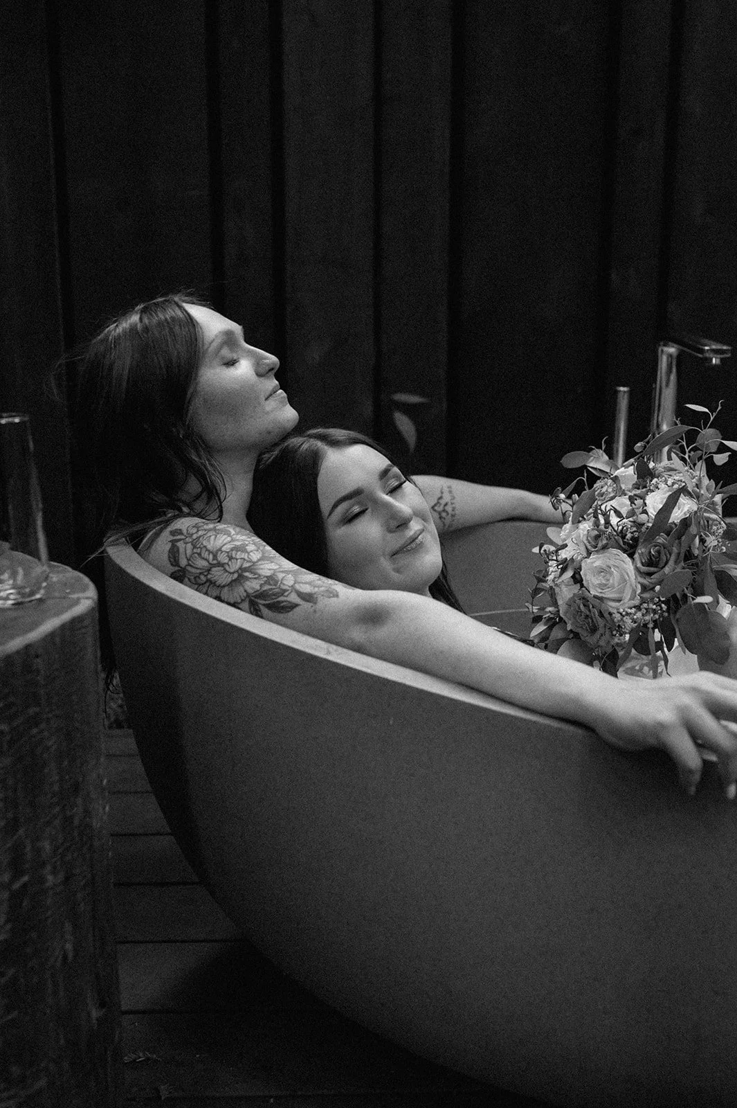 Two women are relaxing together in a bathtub, smiling with closed eyes. One woman has tattoos on her arm and is resting her head against the other woman, who is holding a bouquet of flowers.