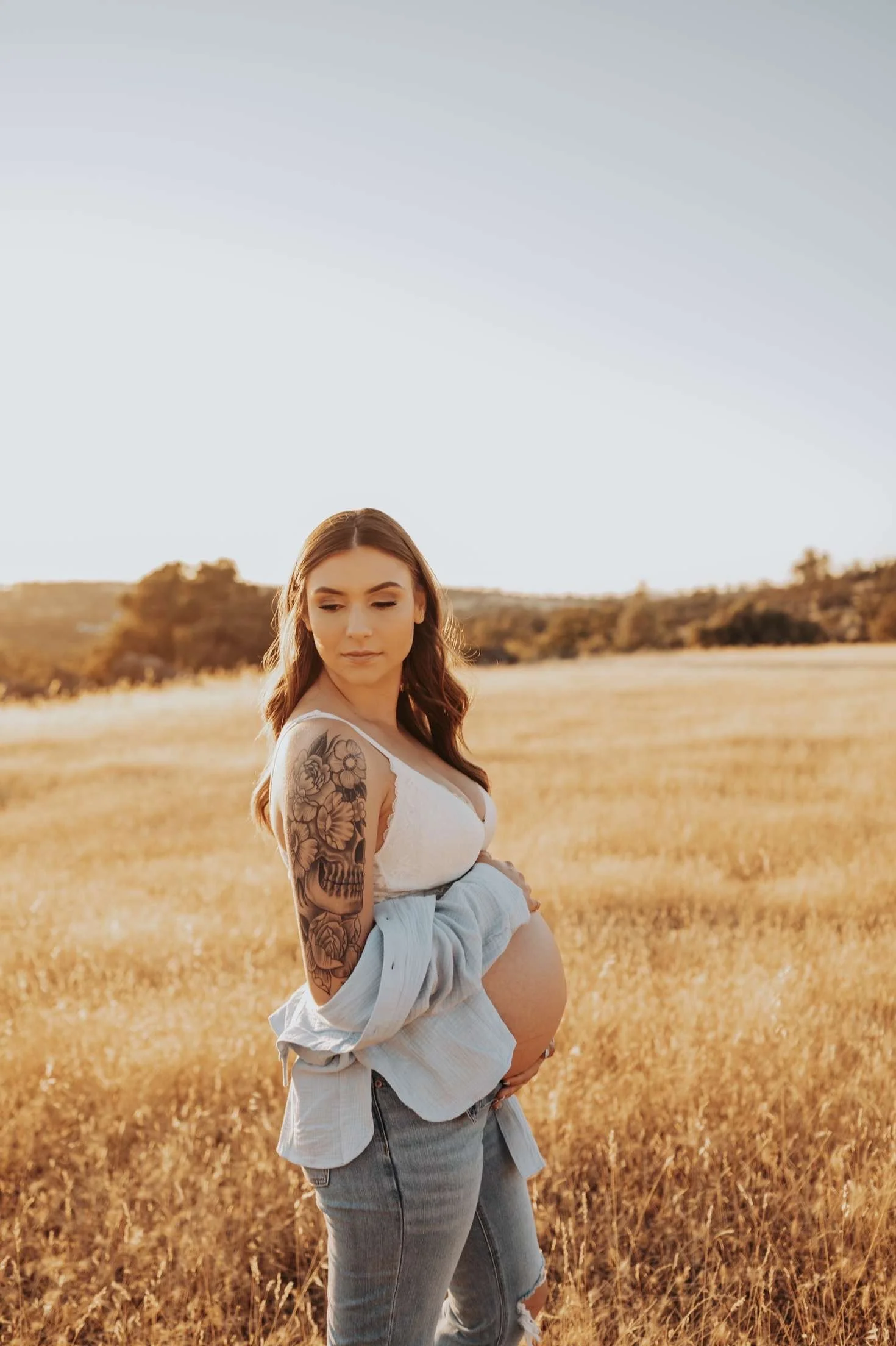 A pregnant woman stands in a golden field during sunset, wearing a white top and ripped jeans, with tattoos on her left arm.