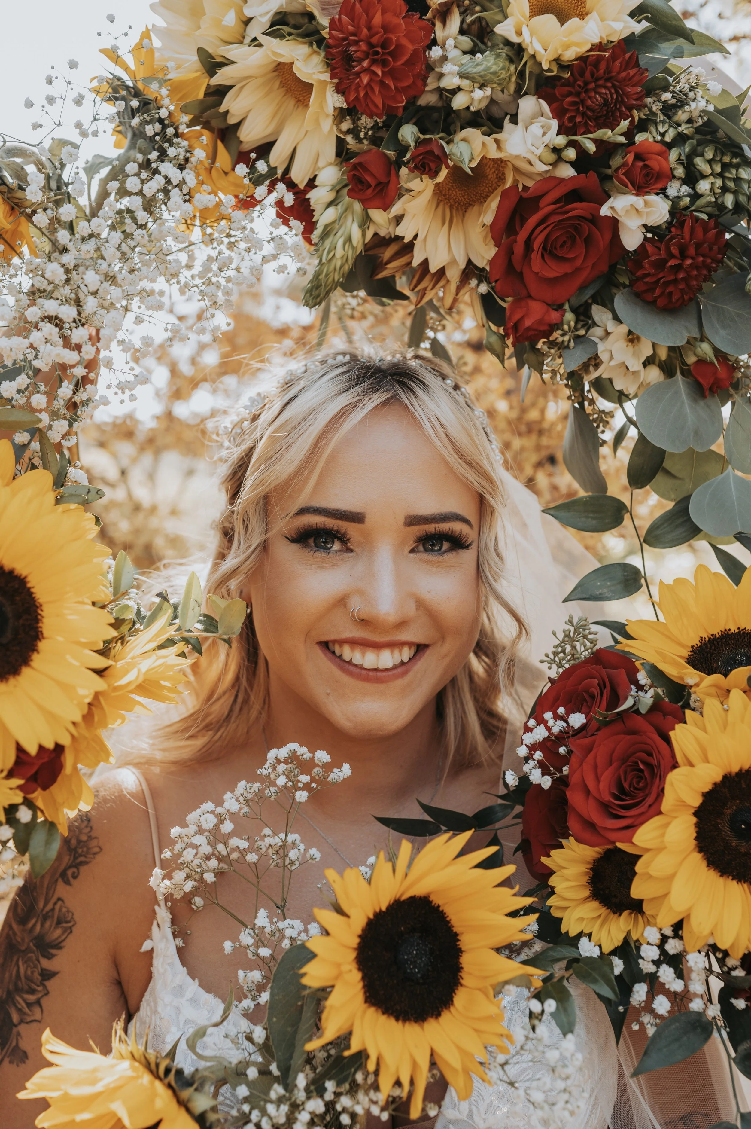A smiling woman with blonde hair, floral makeup, and a nose ring, surrounded by sunflowers, red roses, white baby's breath, and other flowers, outdoors with warm autumn sunlight in the background.