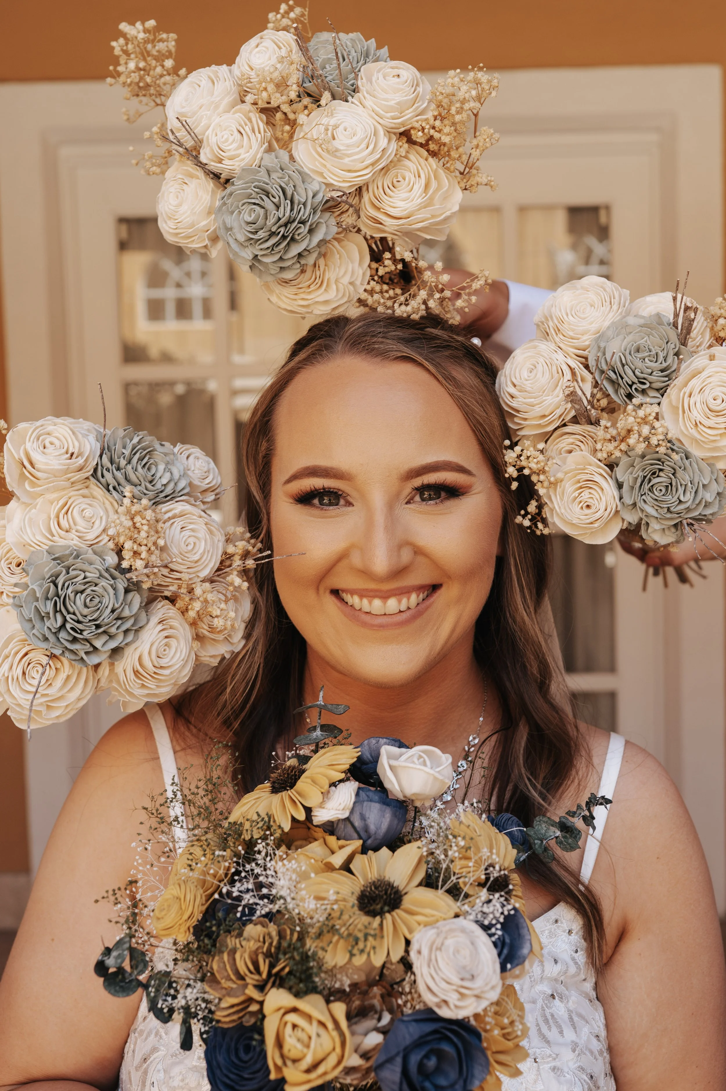 A woman smiling with flowers around her face, holding a bouquet of yellow, white, and blue flowers. Part of another person behind her is holding white and gray flowers above her head.