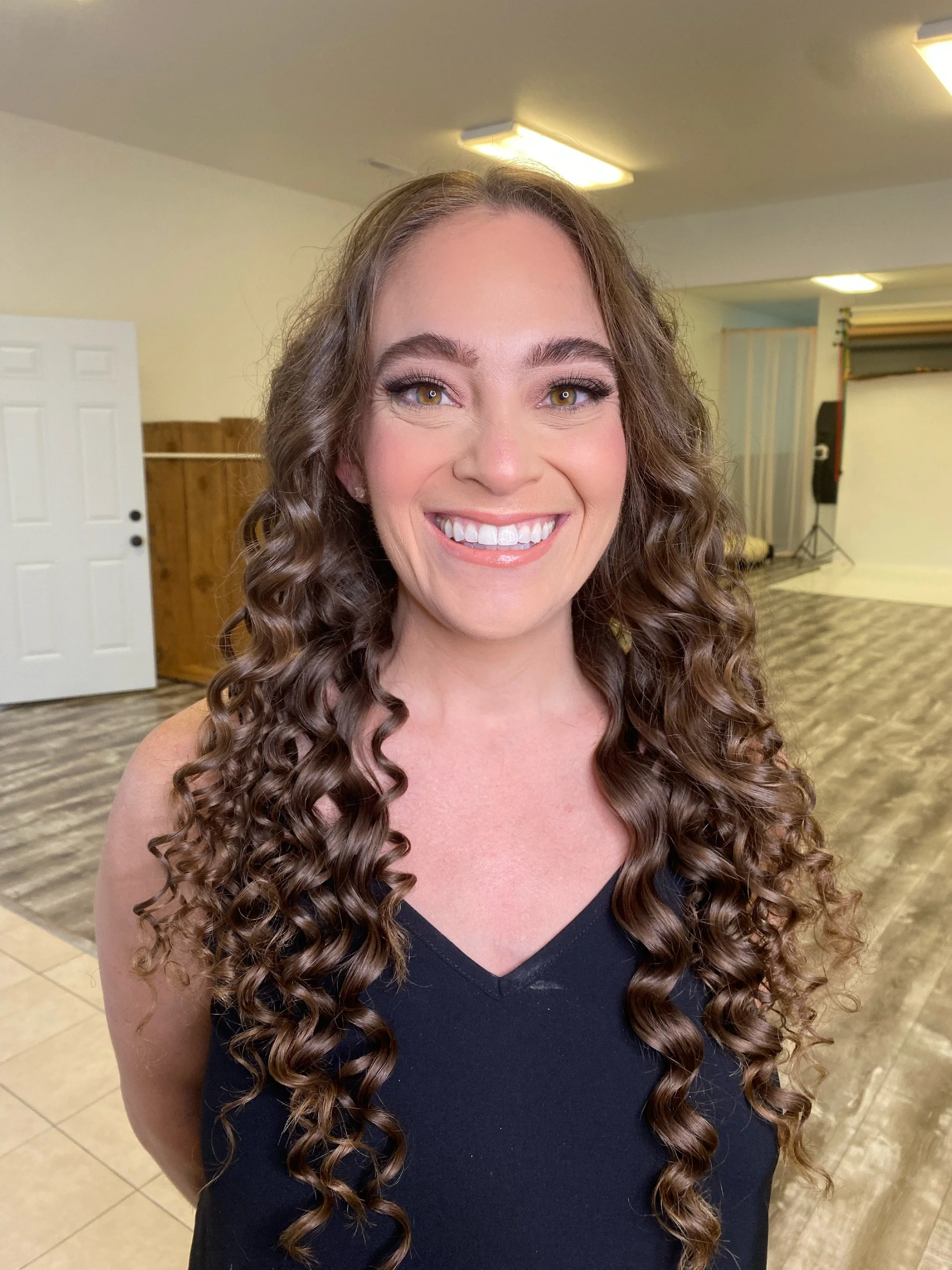 Smiling woman with long curly brown hair in a black top, standing indoors with a background of wooden paneling, a door, and photography equipment.