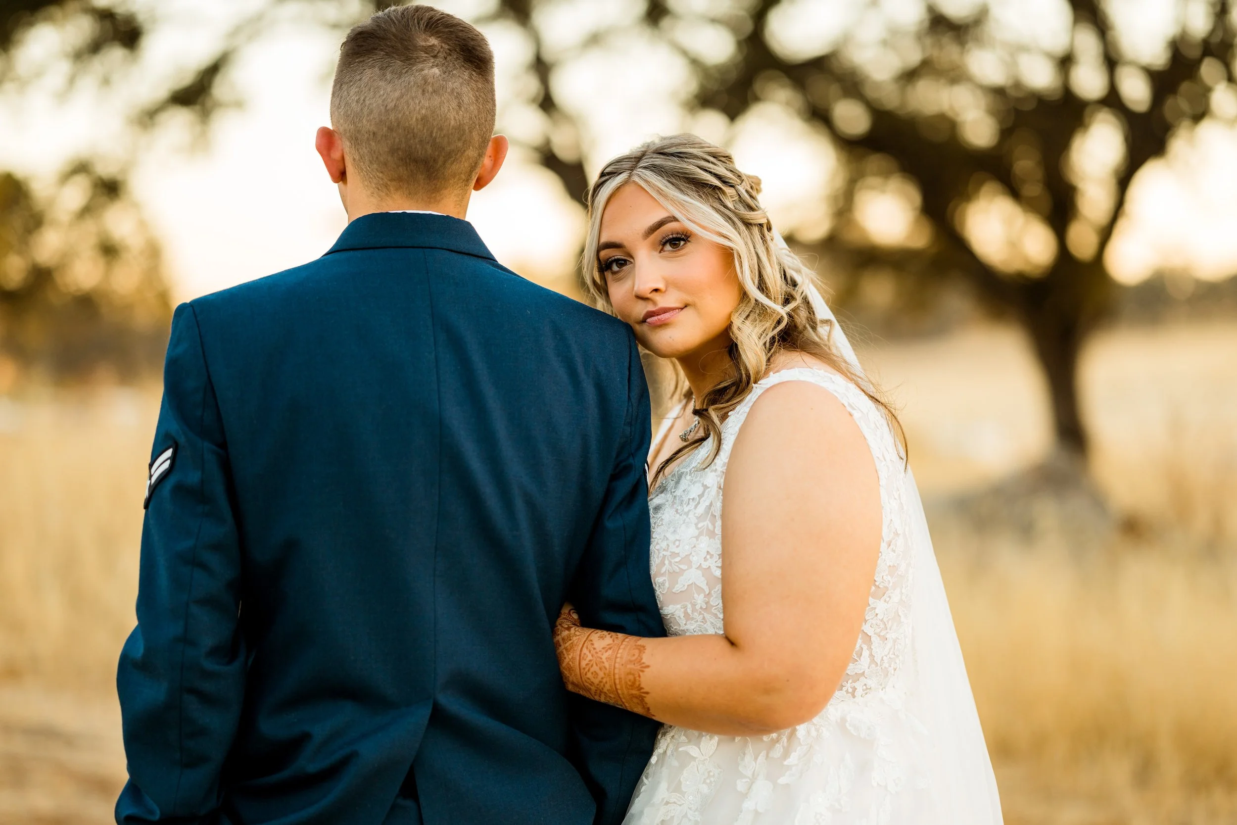 A bride and groom outdoors during sunset, with trees in the background. The bride is looking at the camera with a gentle expression, wearing a lace wedding dress. The groom, dressed in a dark blue suit, has his back to the camera.