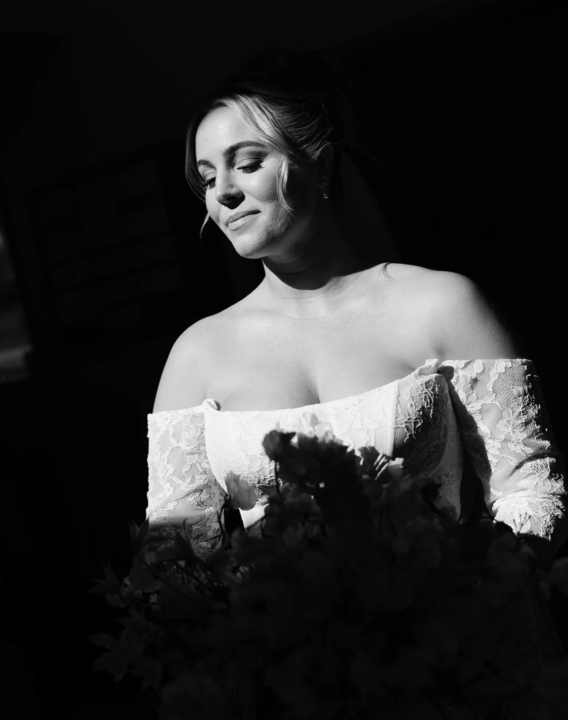 Black and white photo of a bride with closed eyes, wearing an off-shoulder lace wedding dress, holding a bouquet of flowers, with soft lighting highlighting her face and upper body.