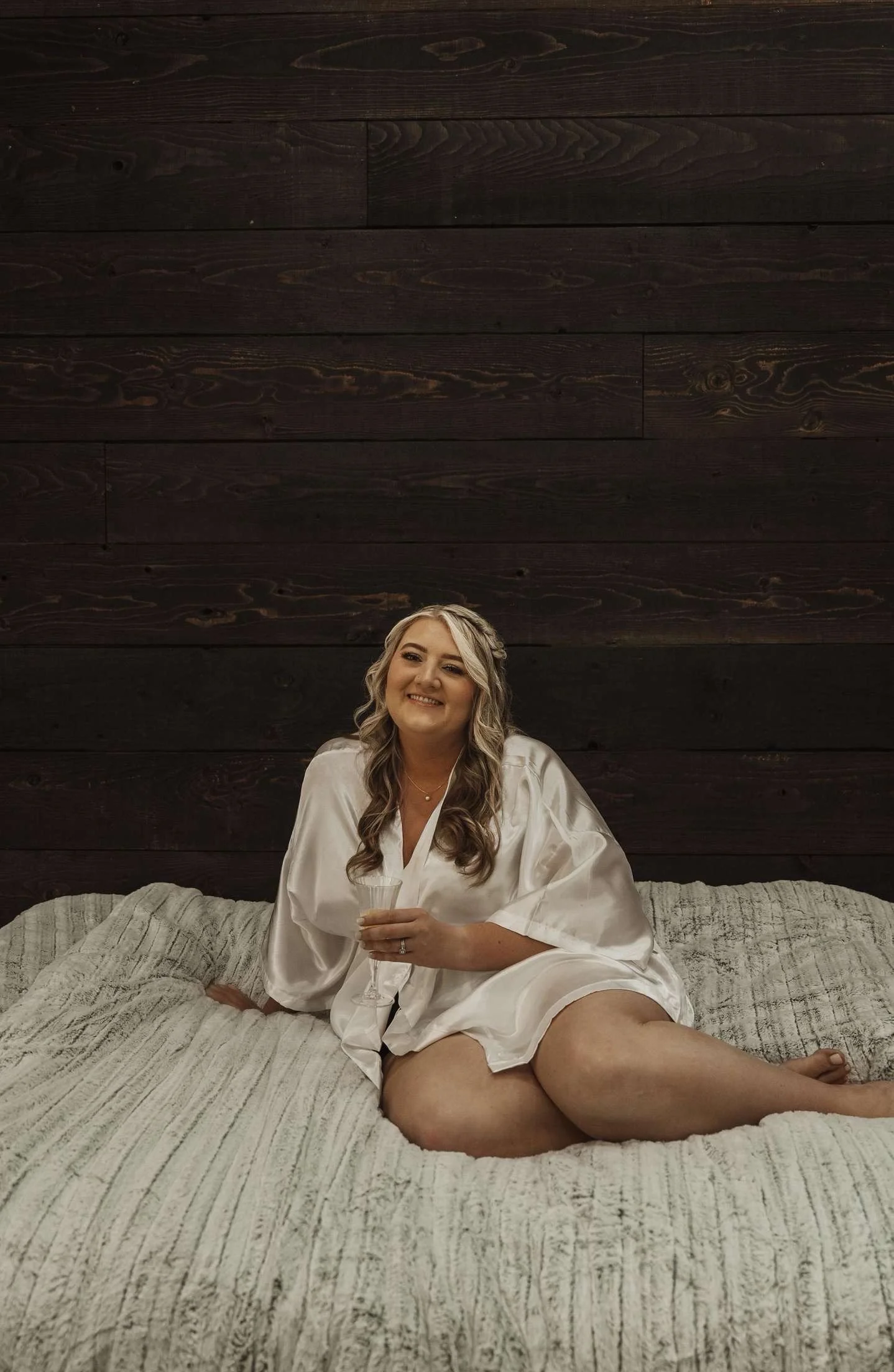 A woman with blonde hair sitting on a bed, smiling, holding a glass of champagne, wearing a white silk robe