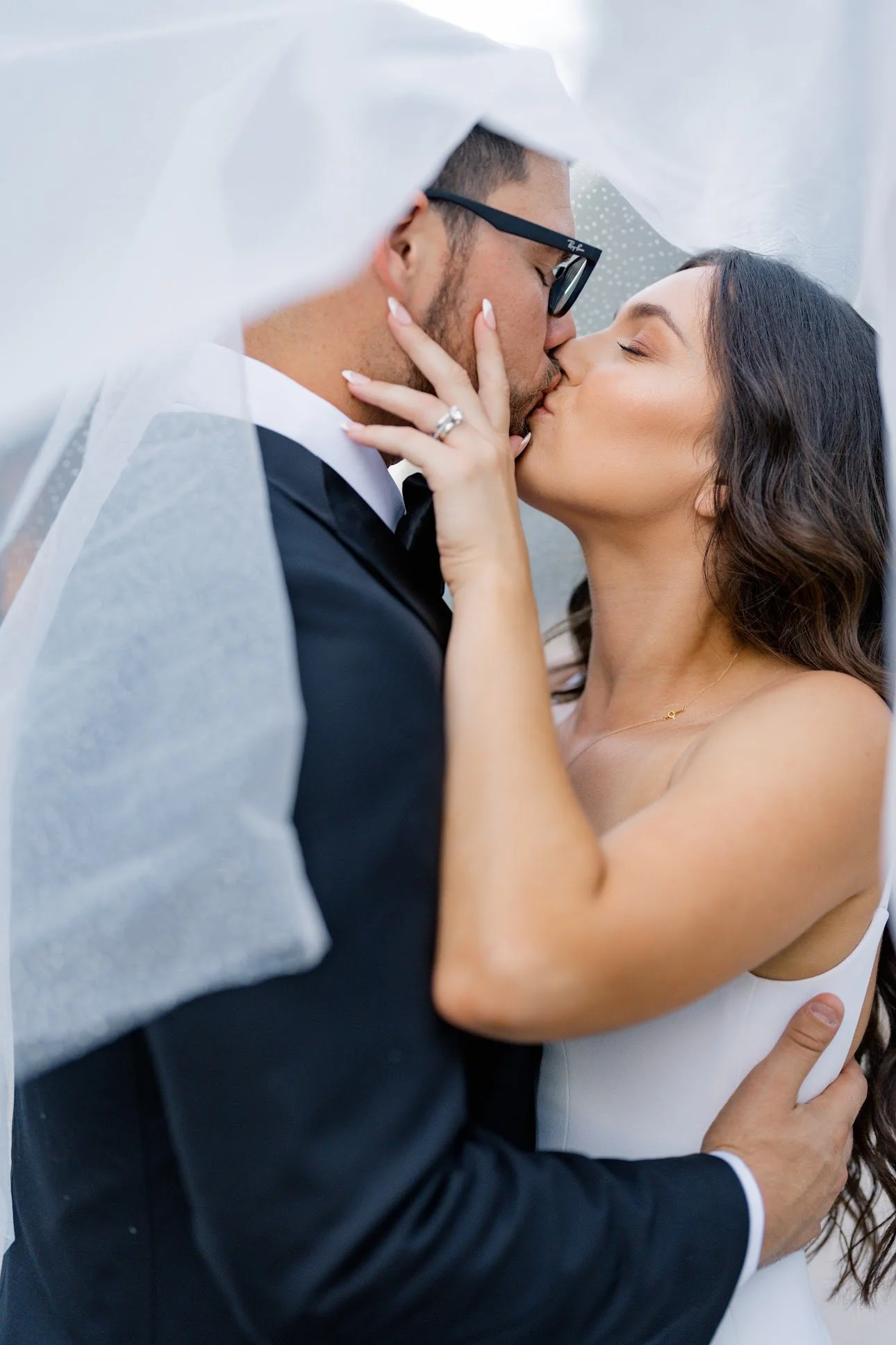 A bride and groom sharing a kiss under a white veil outdoors.