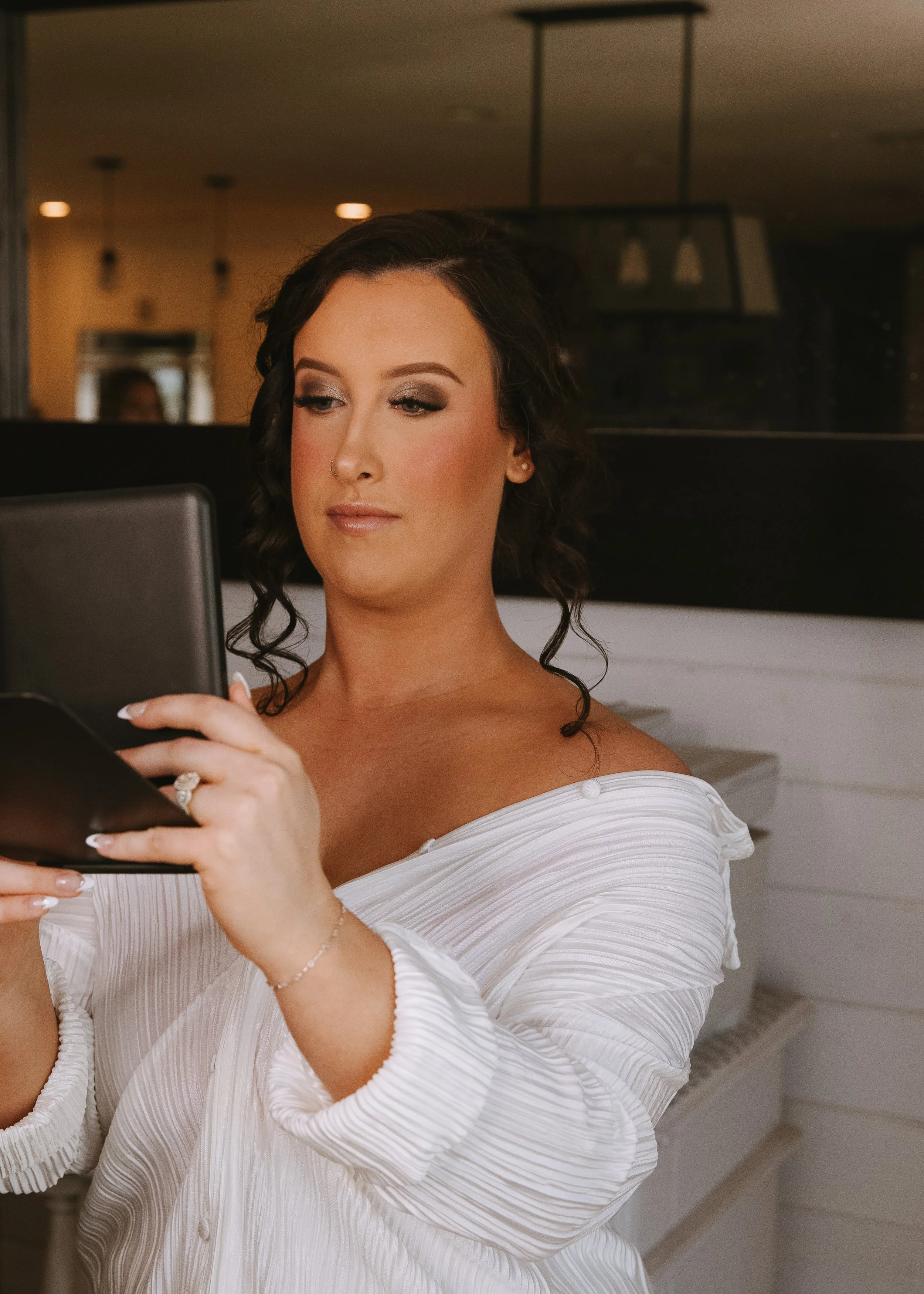 A woman with dark hair styled in loose curls, wearing makeup and a white off-the-shoulder dress, is looking at her phone indoors.