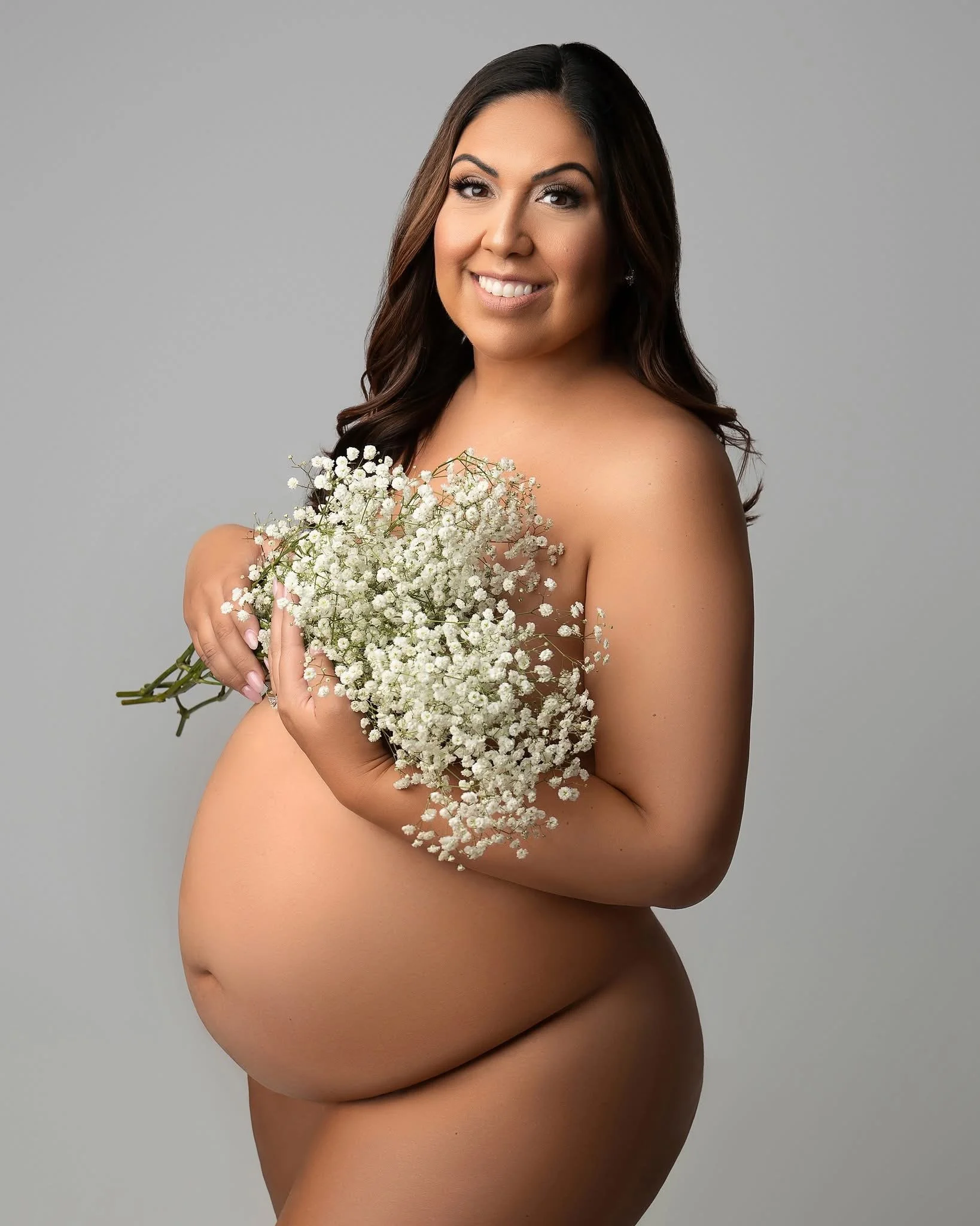 Nude woman holding a bouquet of white baby's breath flowers, smiling, against a plain gray background.