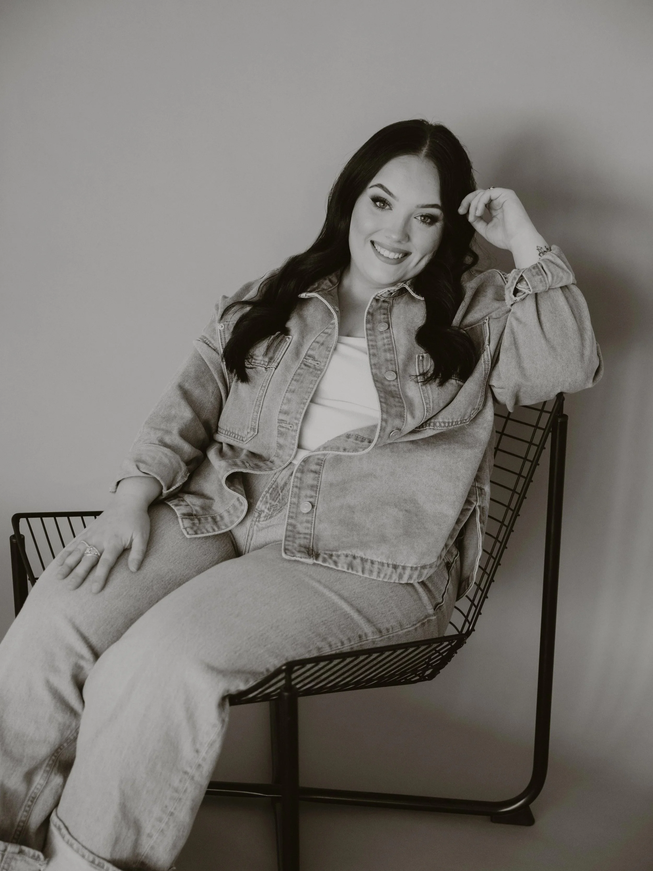 Black and white photo of a smiling woman with dark hair, wearing a denim jacket, sitting in a wire chair against a plain wall.