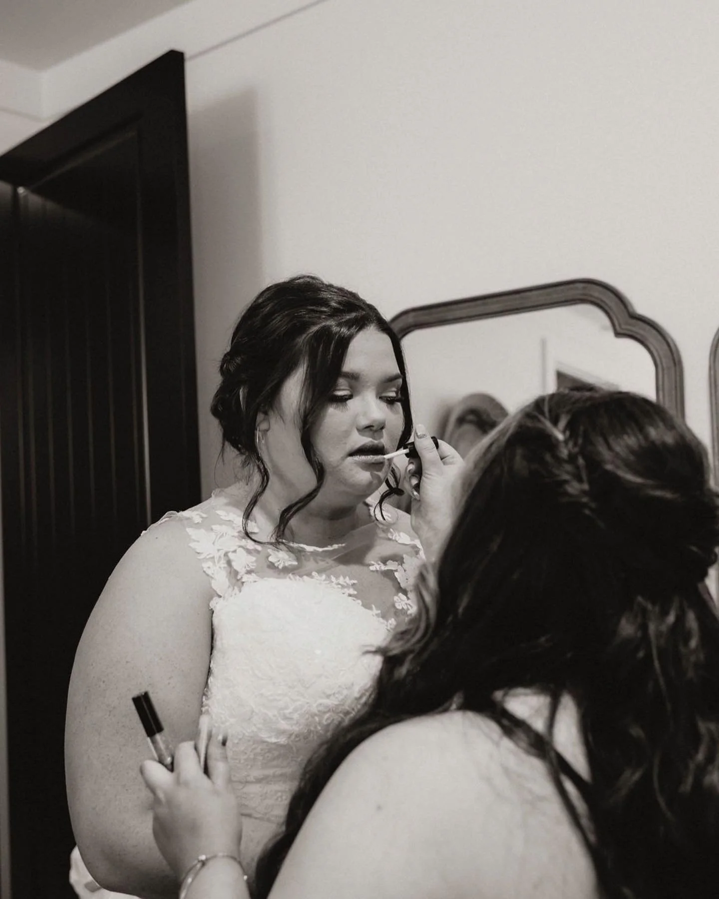 A woman in a wedding dress having her makeup done by another woman in front of a mirror.
