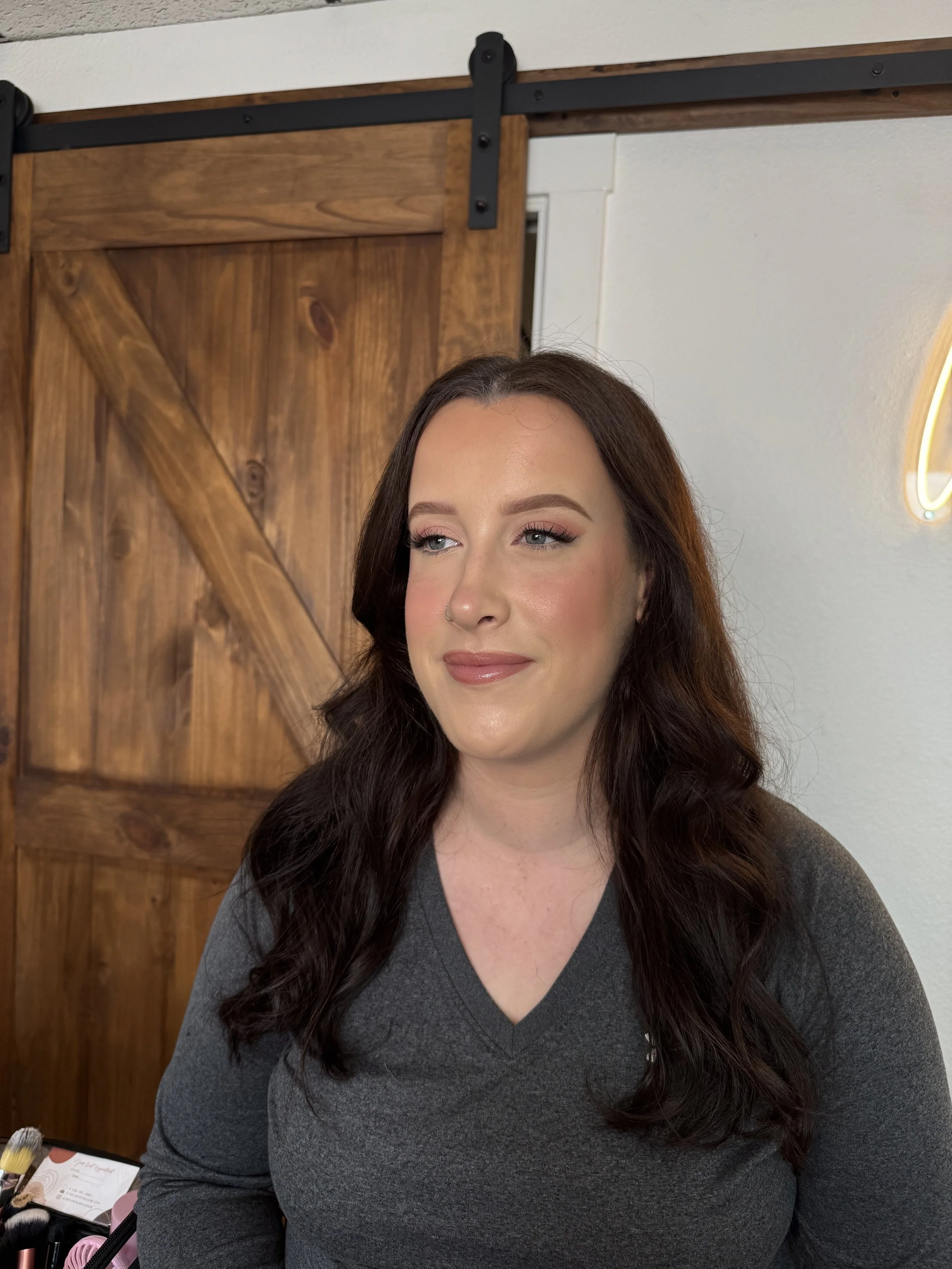 A woman with dark brown wavy hair and makeup, wearing a gray shirt, looking slightly to the side, standing indoors with a wooden sliding barn door and a white wall in the background.