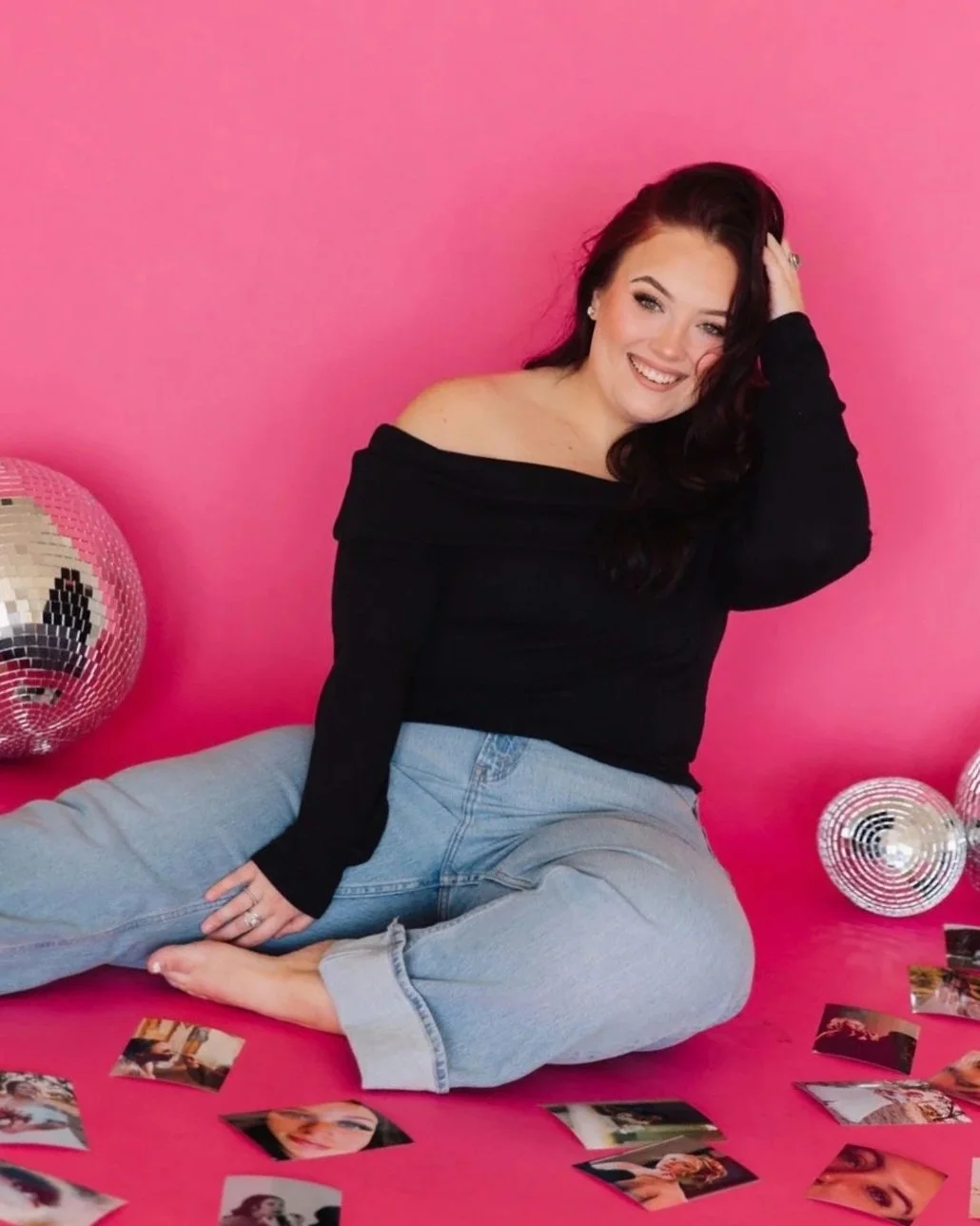 A young woman with dark hair, wearing a black off-the-shoulder top and light blue jeans, sitting on a pink floor in front of a pink wall. She is smiling and touching her hair, surrounded by disco balls and scattered photographs.