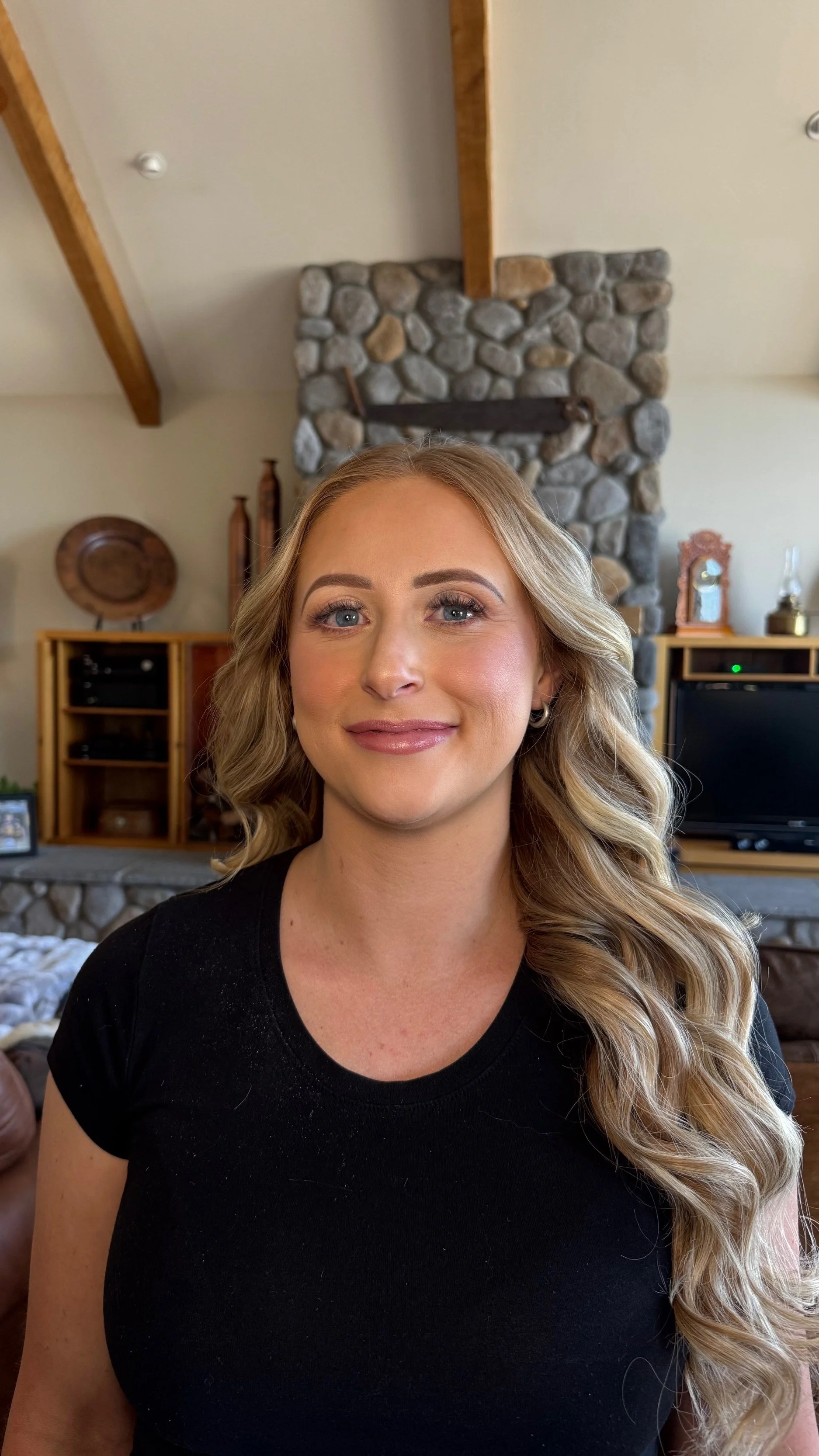 A woman with blonde, wavy hair and blue eyes, smiling in a cozy living room with a stone fireplace, wooden shelves, and a TV in the background.