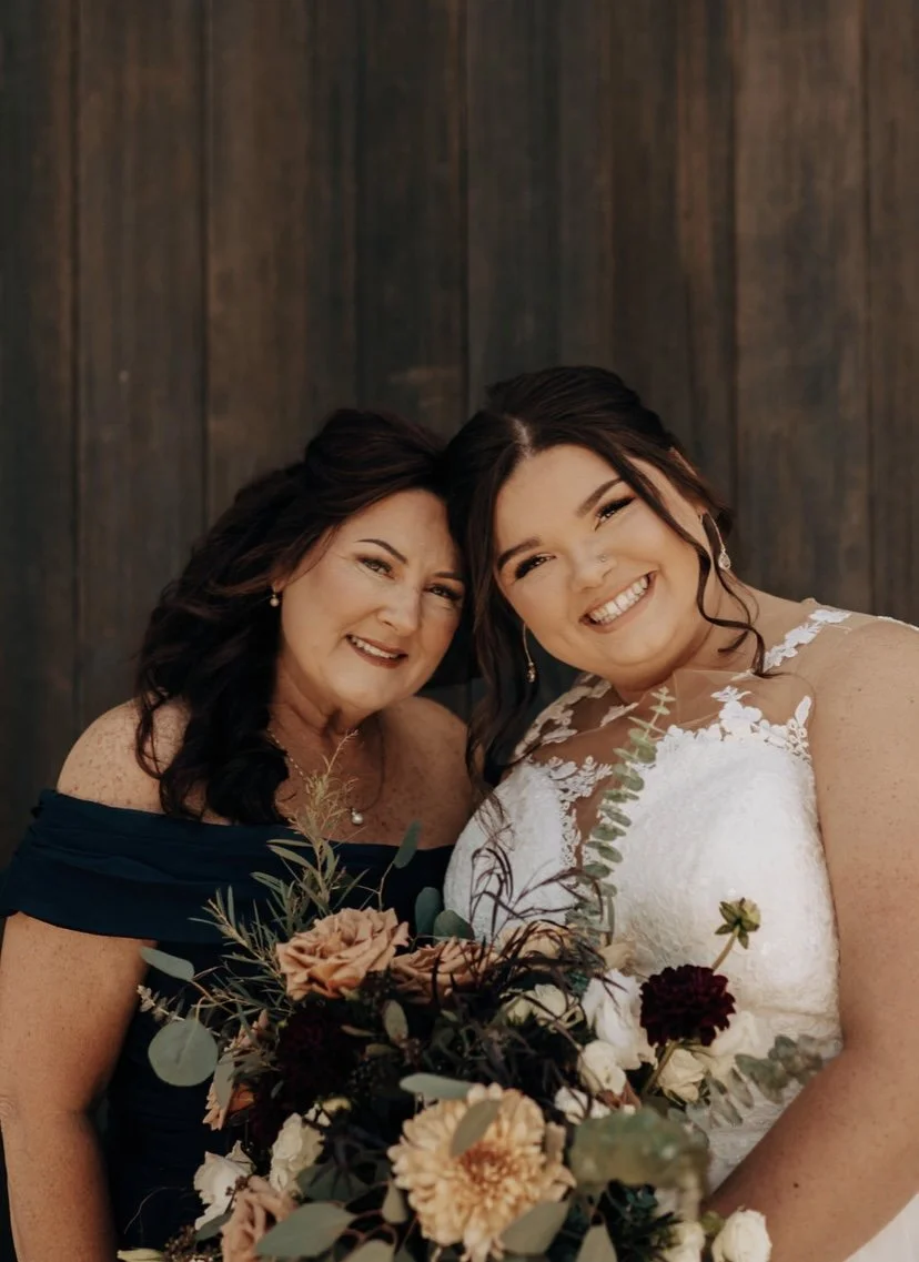 Two women, one older and one younger, smiling and embracing each other at a wedding. The younger woman is in a white lace wedding dress holding a bouquet of flowers. The older woman is in an off-the-shoulder navy blue dress, and they are standing in