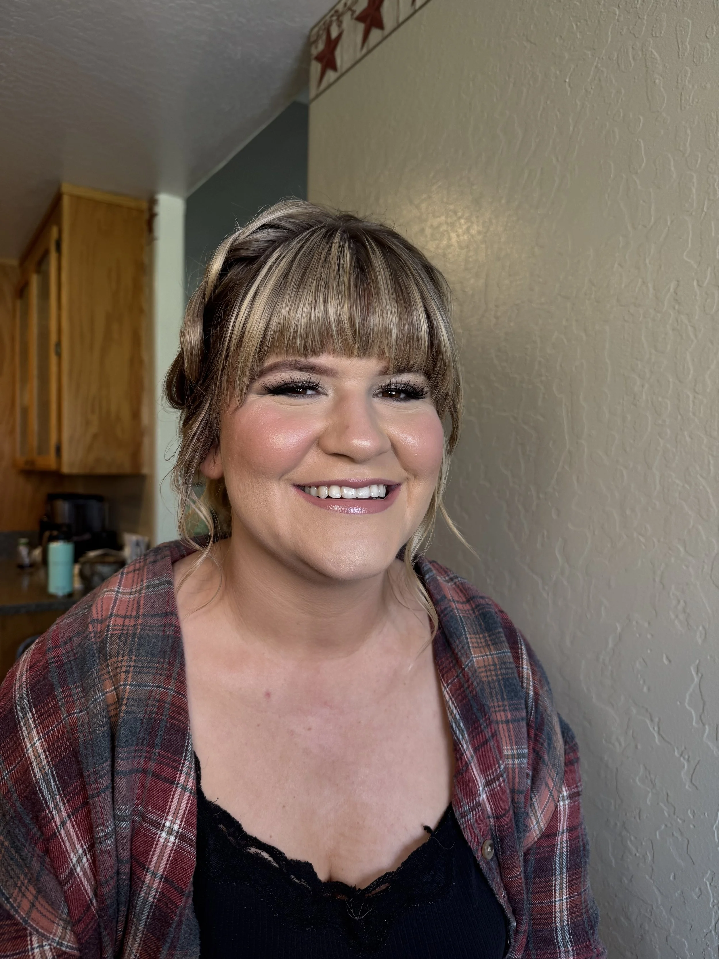 A woman smiling with short blonde hair, wearing makeup, a black top, and a plaid shirt, standing indoors in front of a beige textured wall.