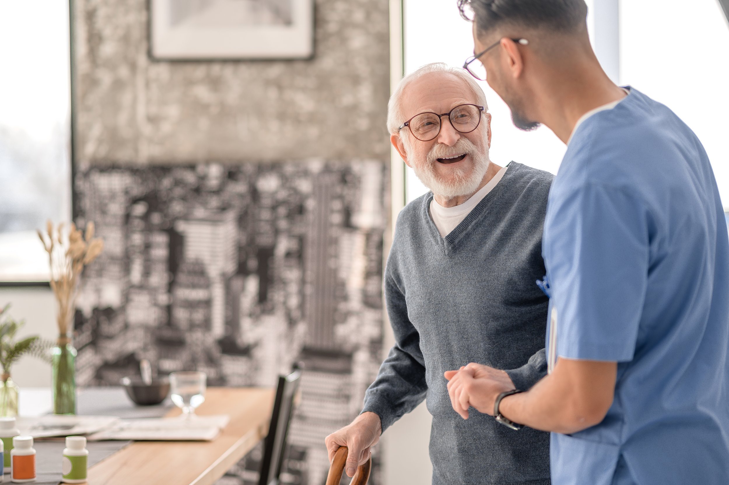 An elderly man with glasses and a gray sweater smiling and talking with a young male healthcare professional in a blue uniform in a bright room.