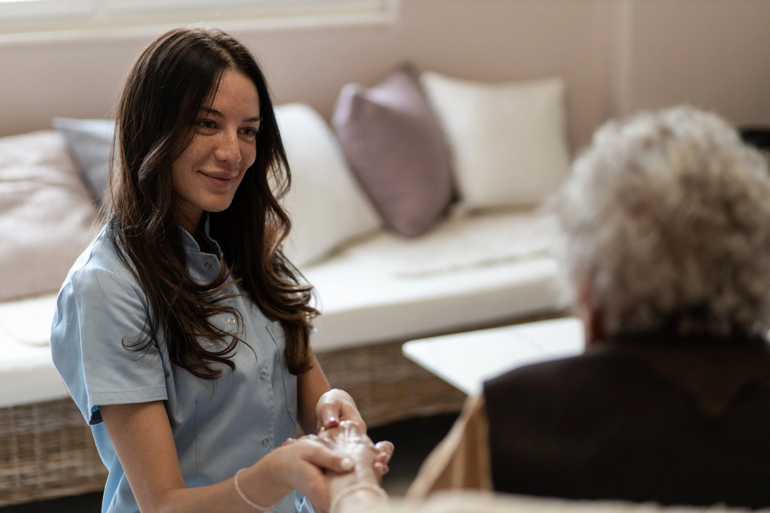 A young woman with long brown hair, wearing a light blue shirt, holding and smiling at an elderly woman with curly white hair, who is partially visible and facing away. They are in a cozy room with a white couch and pillows in the background.