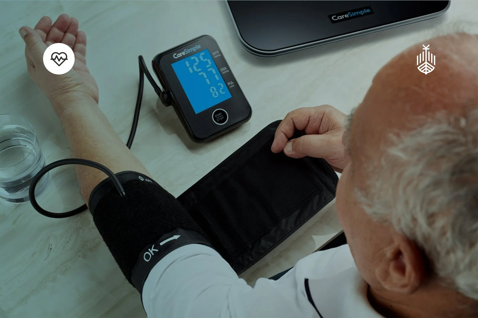Elderly man checking his blood pressure with a digital monitor using a cuff on his arm, on a light-colored table.