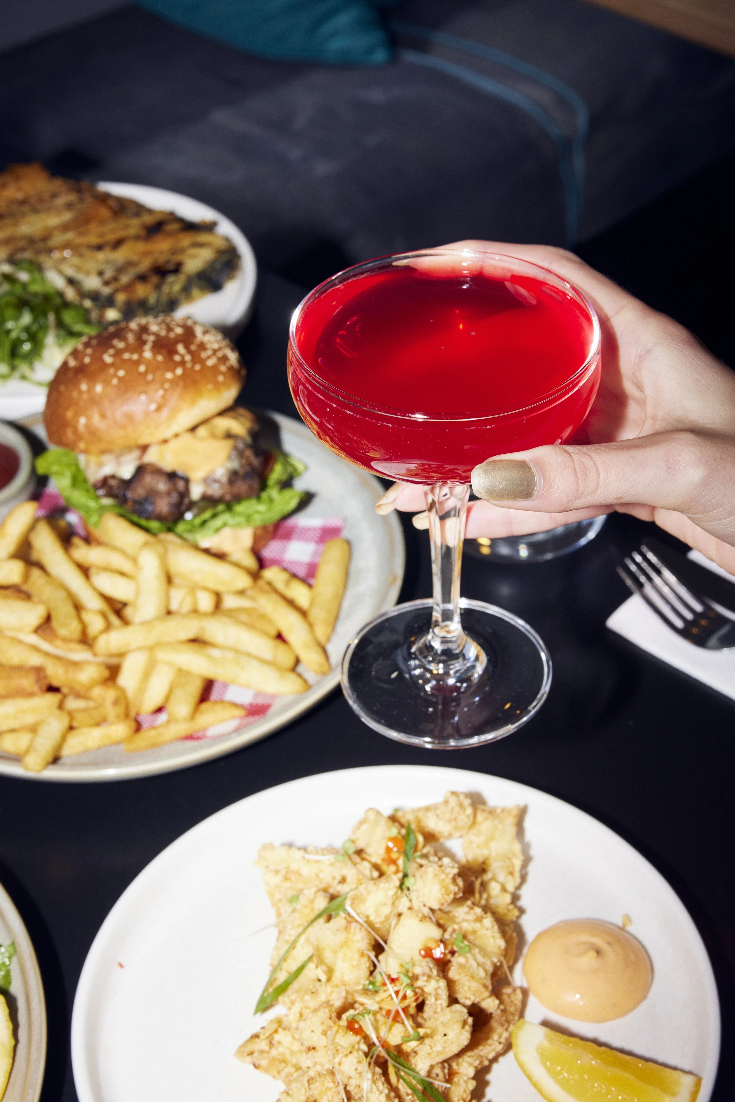 Hand holding a red cocktail beside a gourmet burger and fries on a restaurant table.