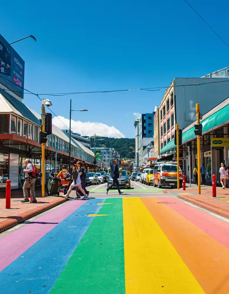 People crossing a vibrant rainbow-painted pedestrian crossing on a sunny day in a busy city street.